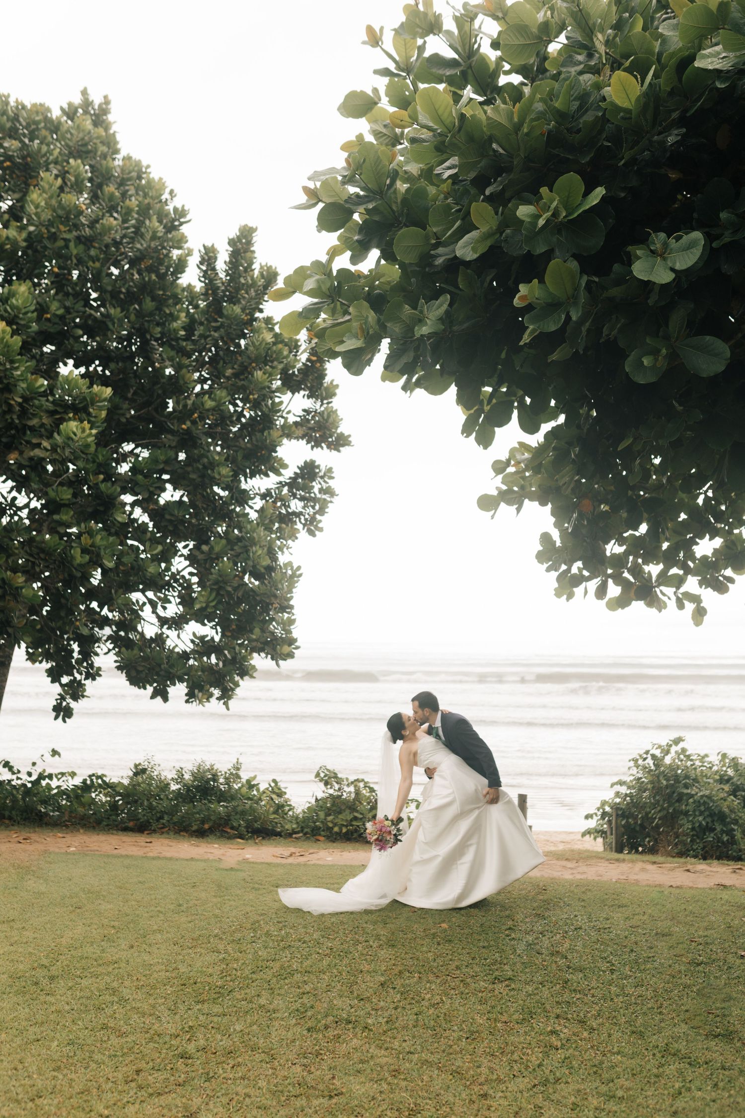 Wedding couple sitting on grass under leafy trees with ocean view.