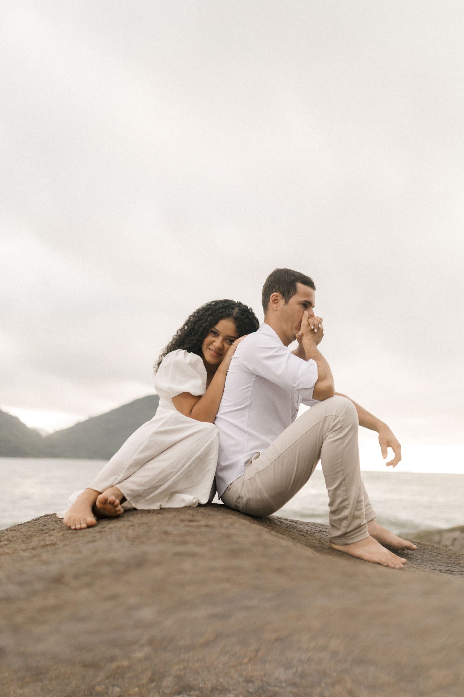 A couple relaxes on a seaside cliff, dressed in white and enjoying the ocean view together.