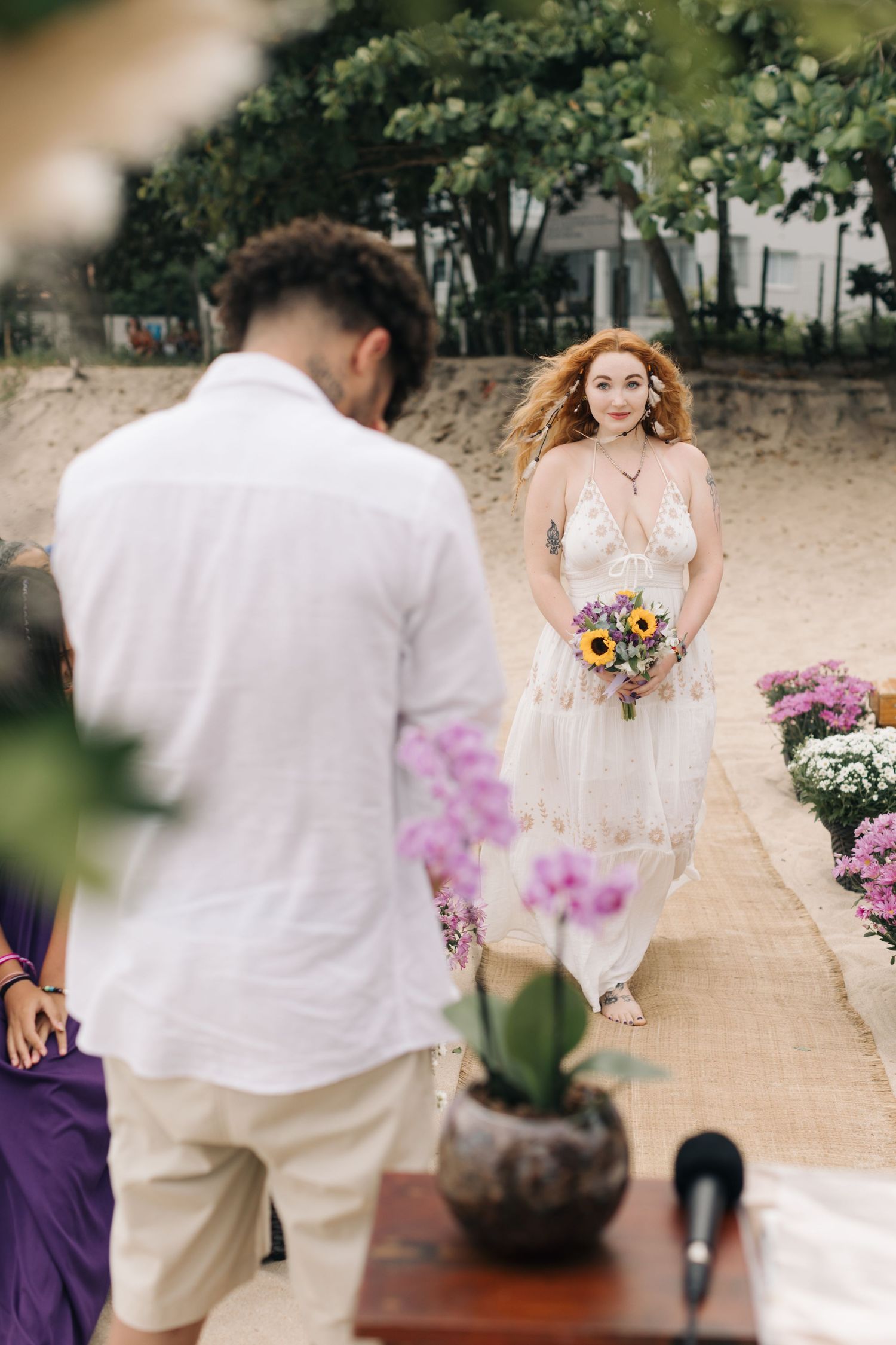 A bride in a delicate white dress carrying a colorful bouquet walks along a beach pathway lined with flowers.
