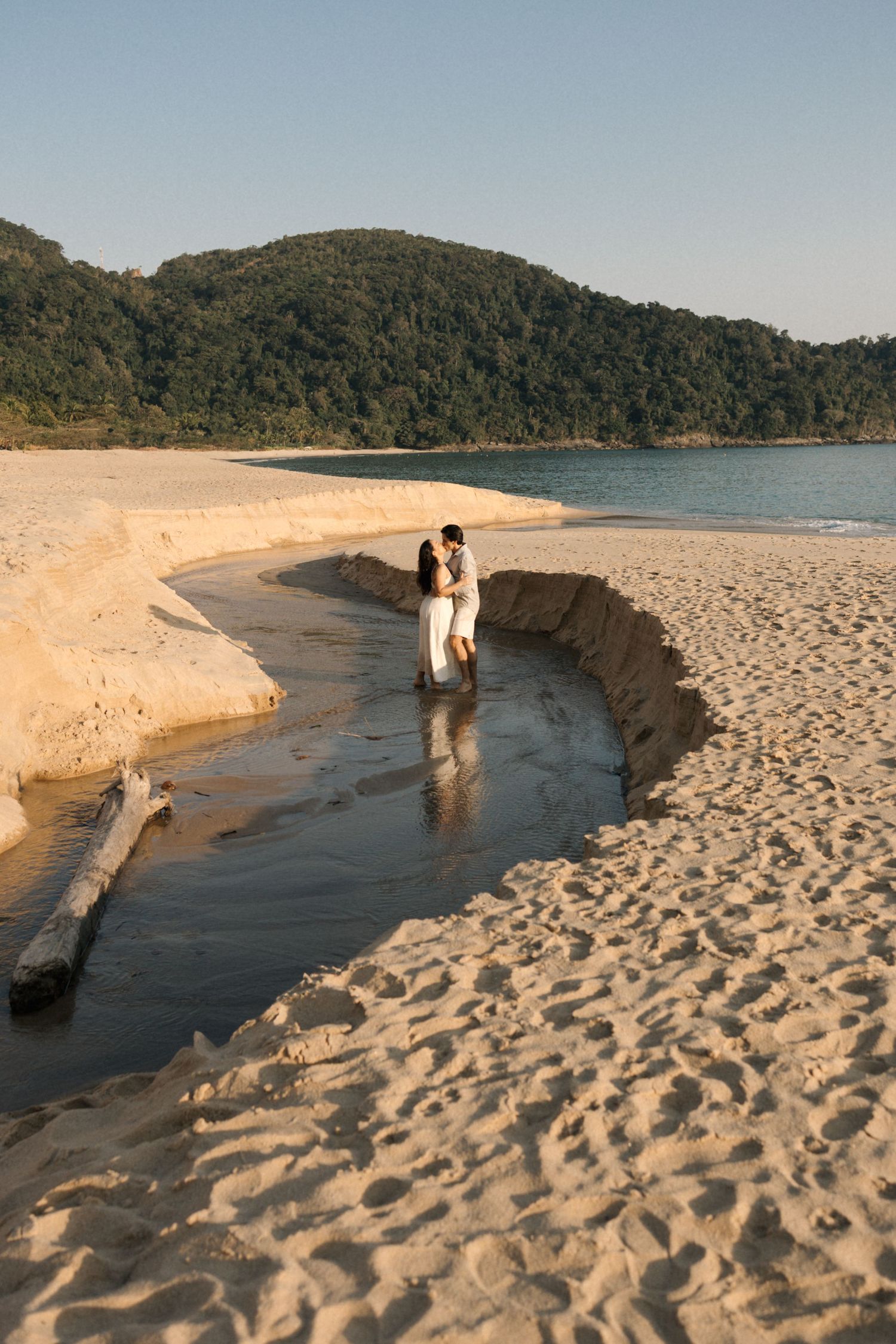 Scenic beach landscape with mountains and a sandy stream leading to the ocean at sunset.