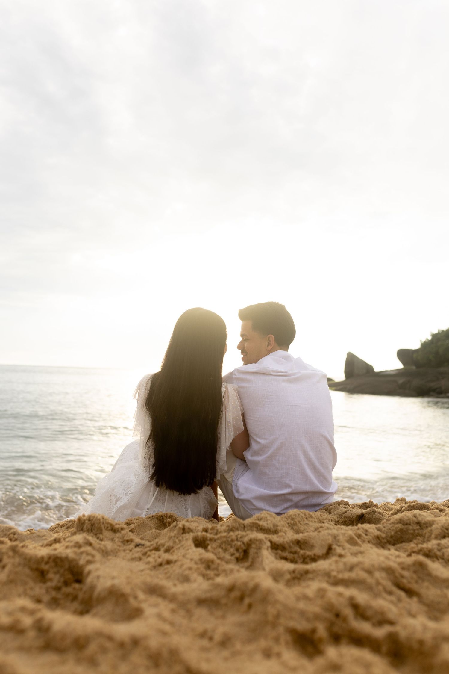 Um casal está sentado em uma praia ao pôr do sol, olhando para o oceano e para a costa rochosa.