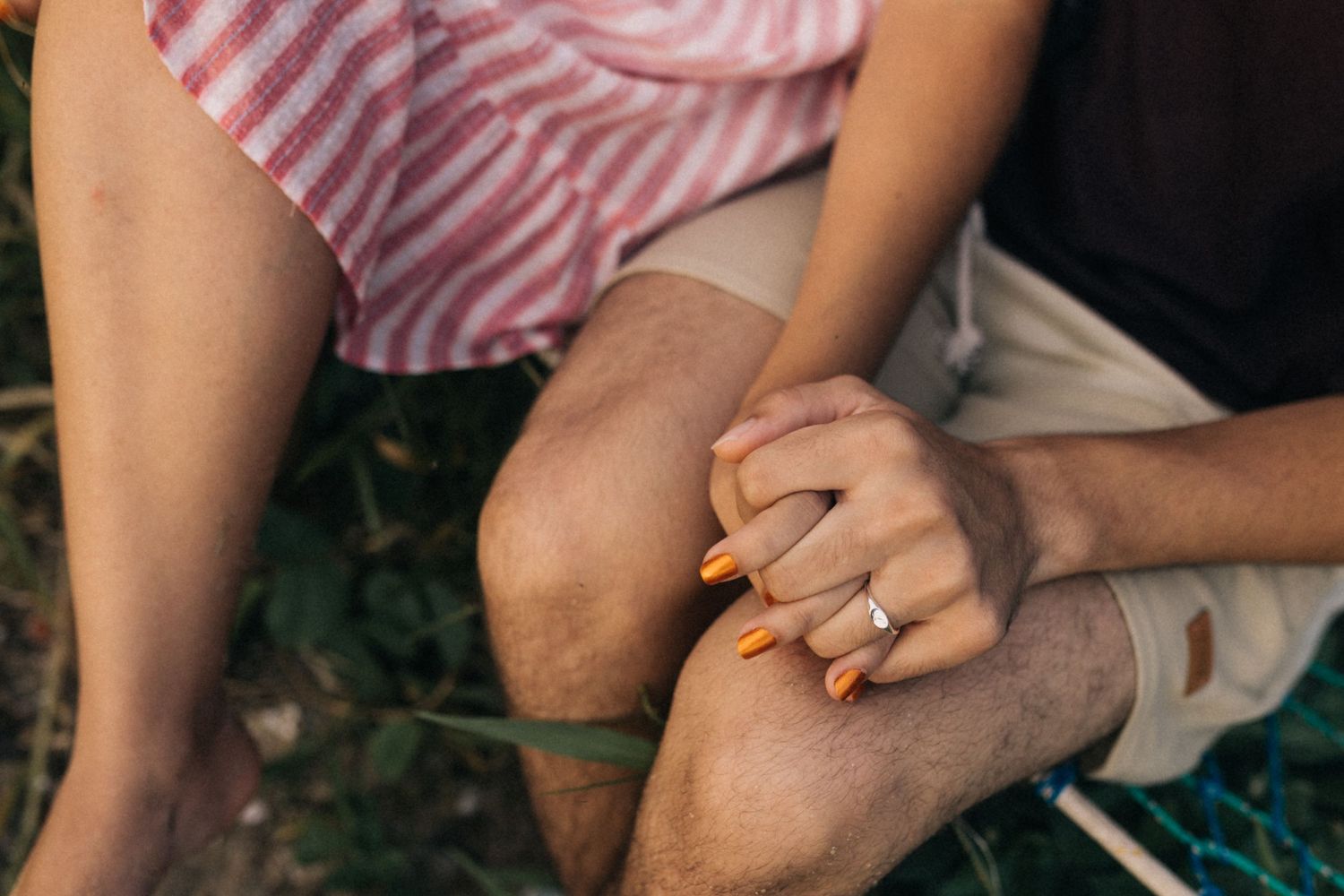 Mãos apoiadas nos joelhos com longas unhas laranja bem cuidadas e roupas listradas rosa e brancas.