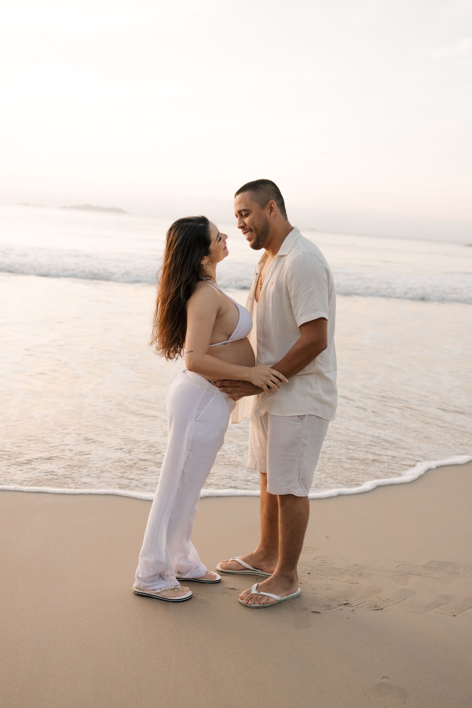 A romantic couple in white attire embraces on a serene beach at sunset.