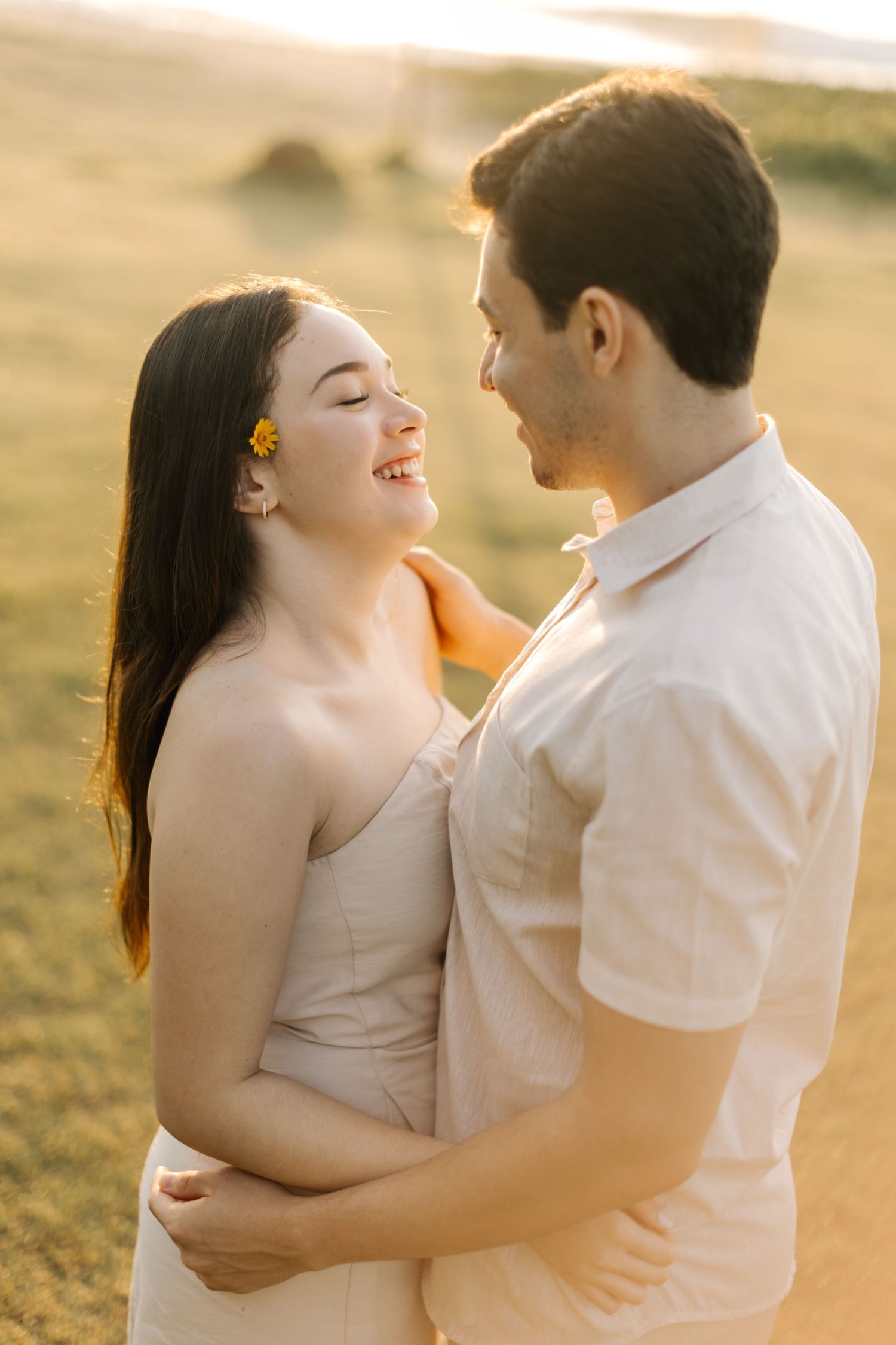 Joyful couple in cream outfits laughing together during sunset photoshoot with golden lighting.