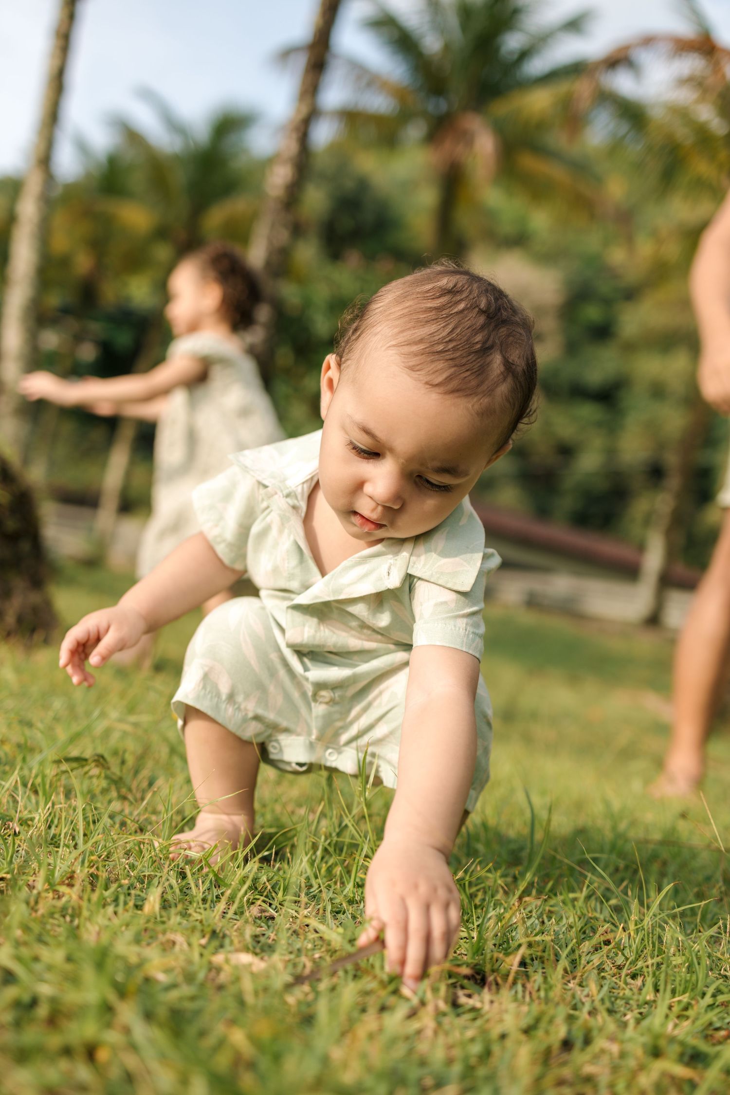 Baby in light colored outfit crawling on grass in a sunny outdoor setting.