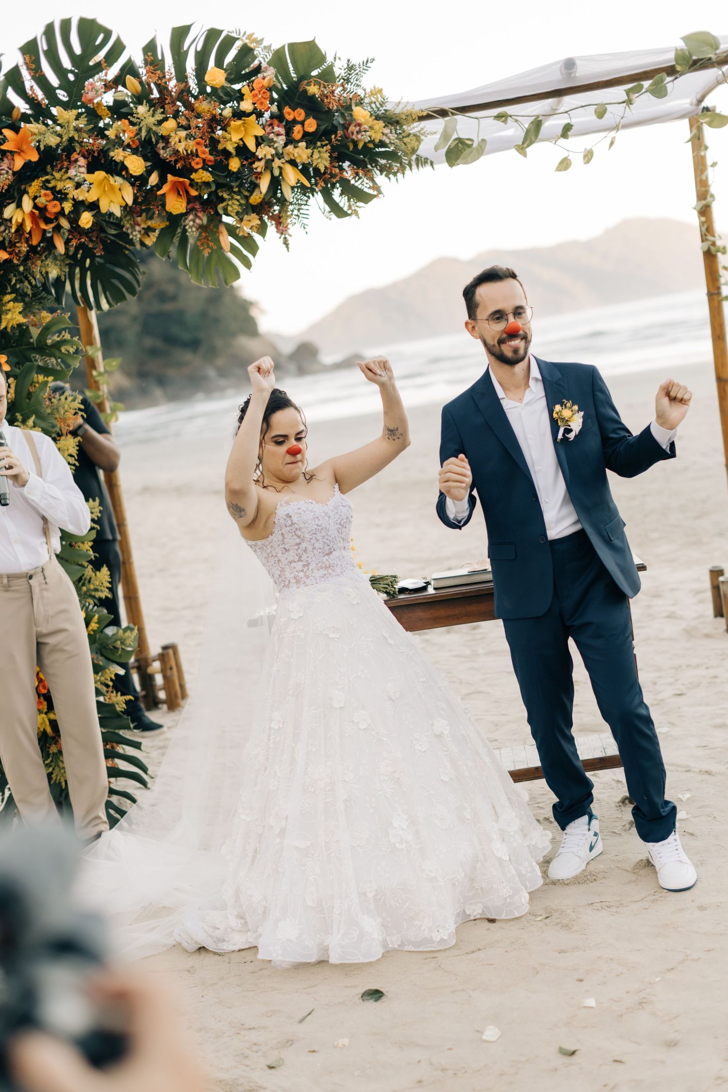 Newlyweds dancing joyfully on beach under tropical floral arch.