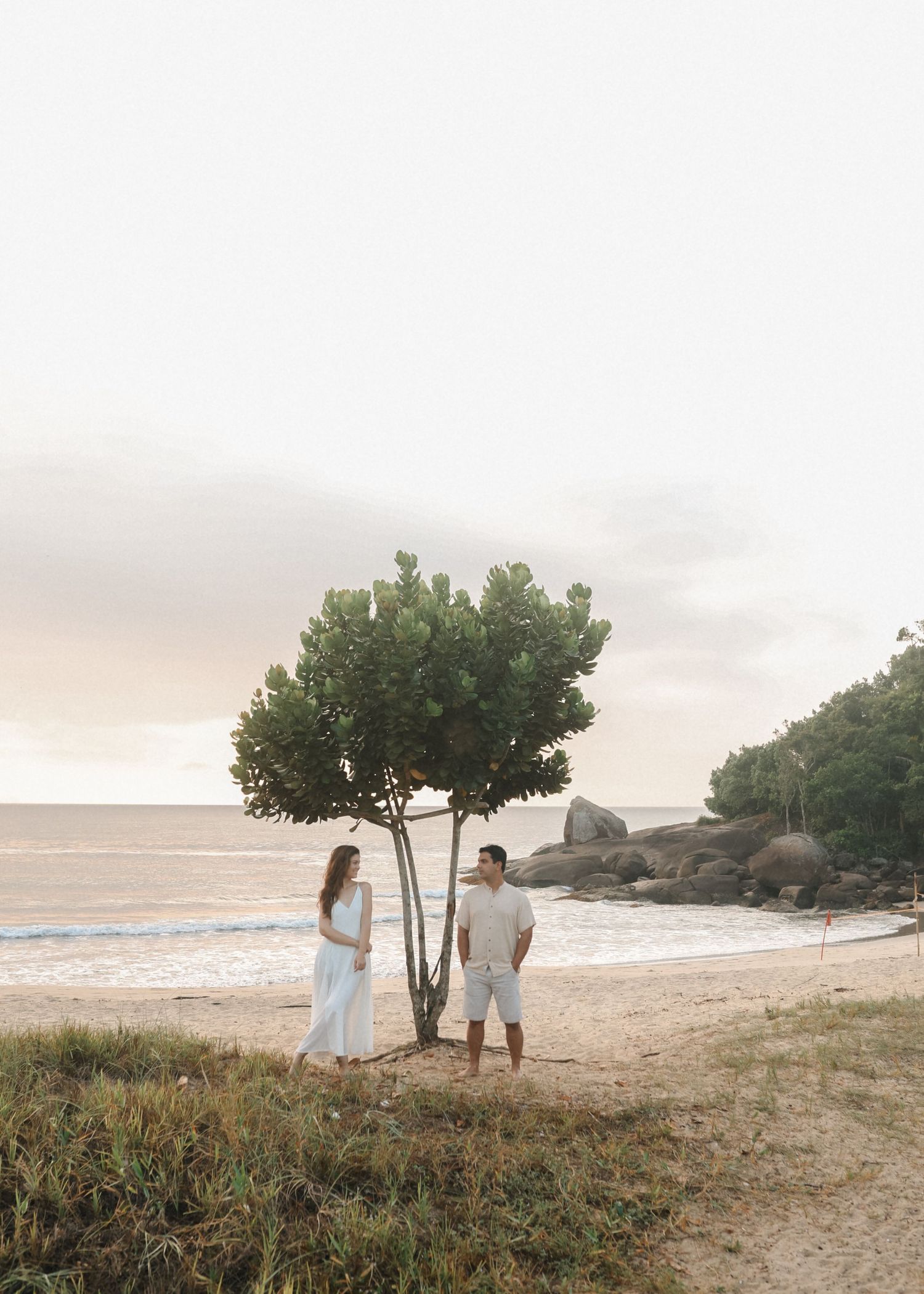 Couple in white walking along scenic beach at sunset with trees in background.