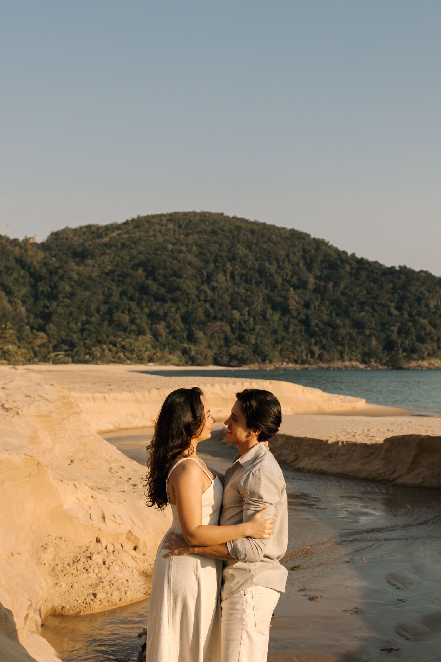 Romantic couple embracing on a sandy beach with green hills in the background during golden hour.