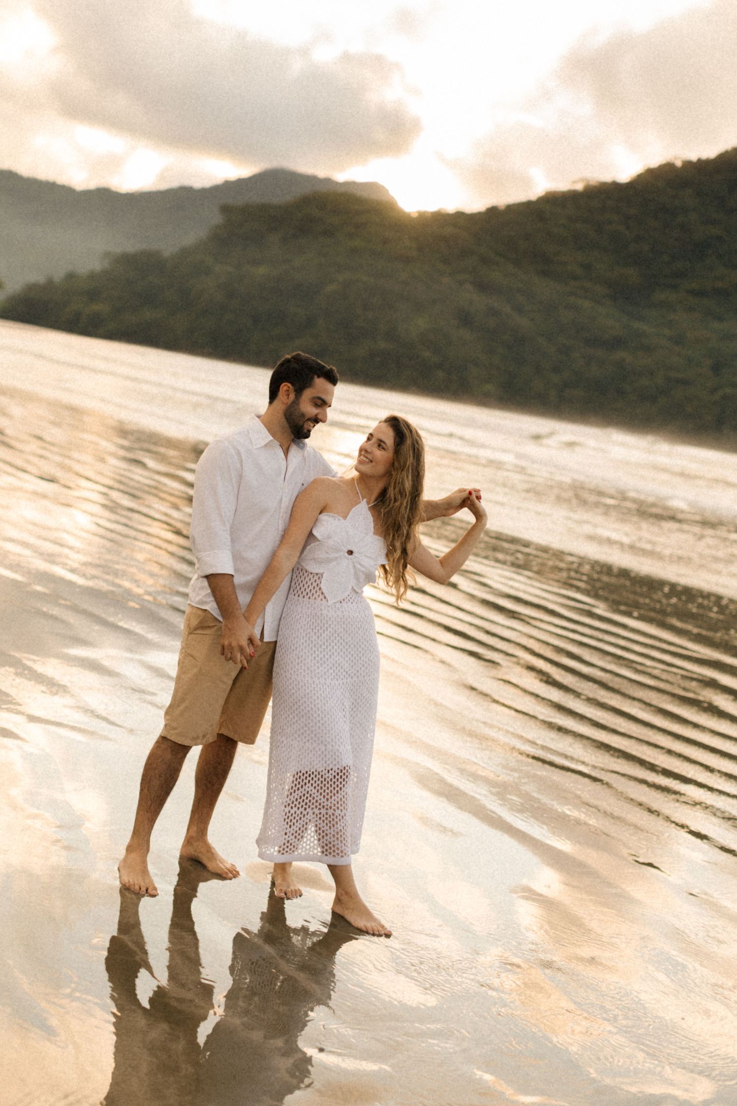 A couple strolls intimately along a scenic beach at sunset with mountains in the background.