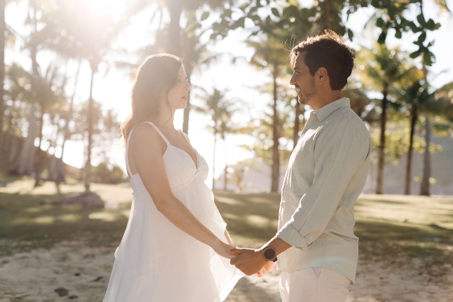 A couple in white and gray outfits holds hands while walking along a sunny beach with palm trees in the background.