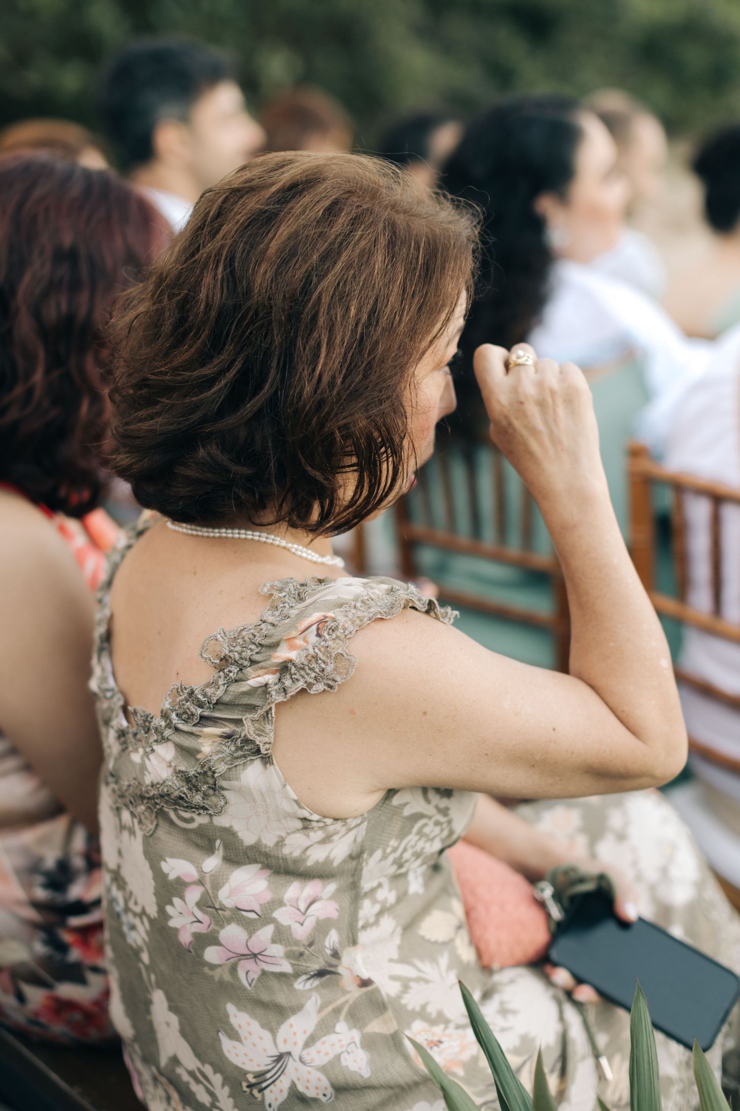 Guest with back tattoo watches wedding ceremony while seated outdoors.