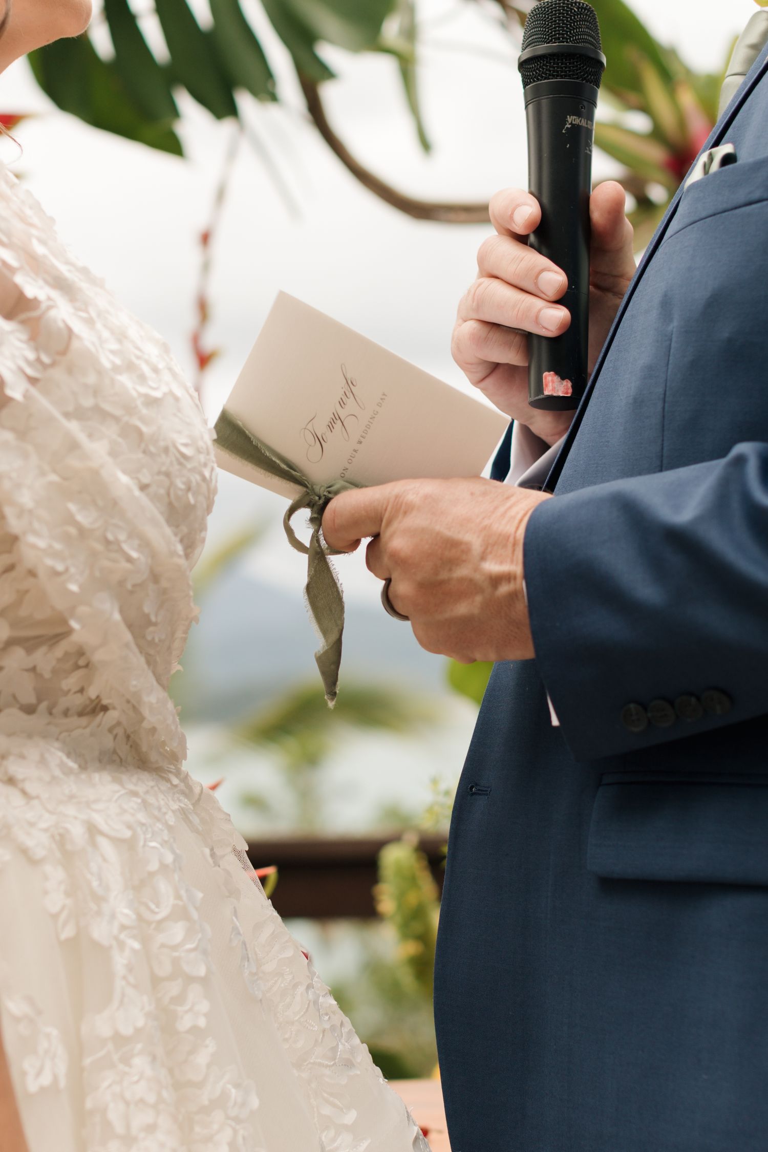 Person in blue suit holding a microphone while standing next to white lace wedding dress details.