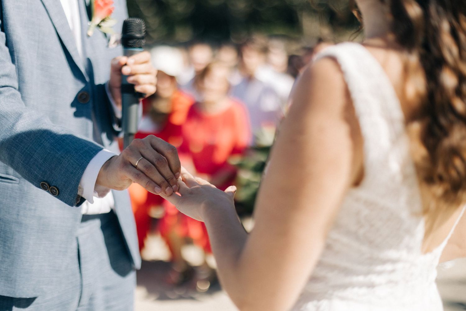 Close-up of couple exchanging rings during outdoor wedding ceremony with guests in background.