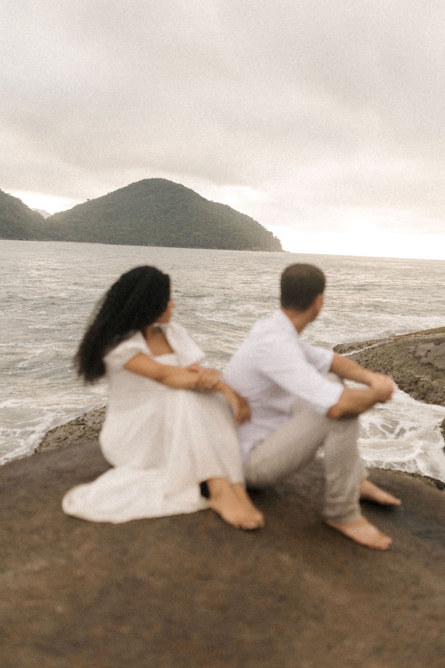 A couple in white clothing sits together on rocky coastal cliffs overlooking the ocean and mountains.