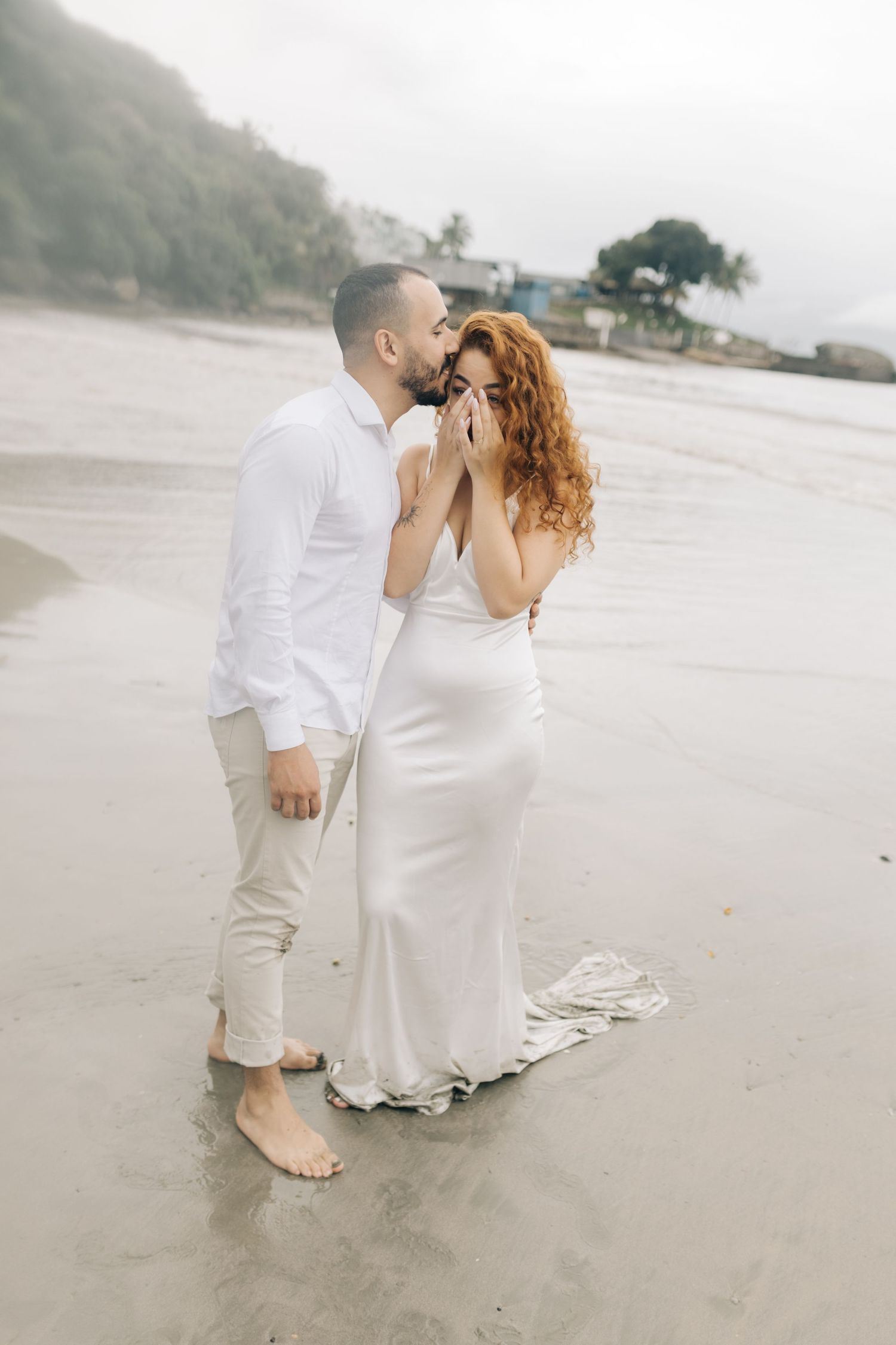 Barefoot couple sharing intimate moment on wet beach sand with mountains in background.