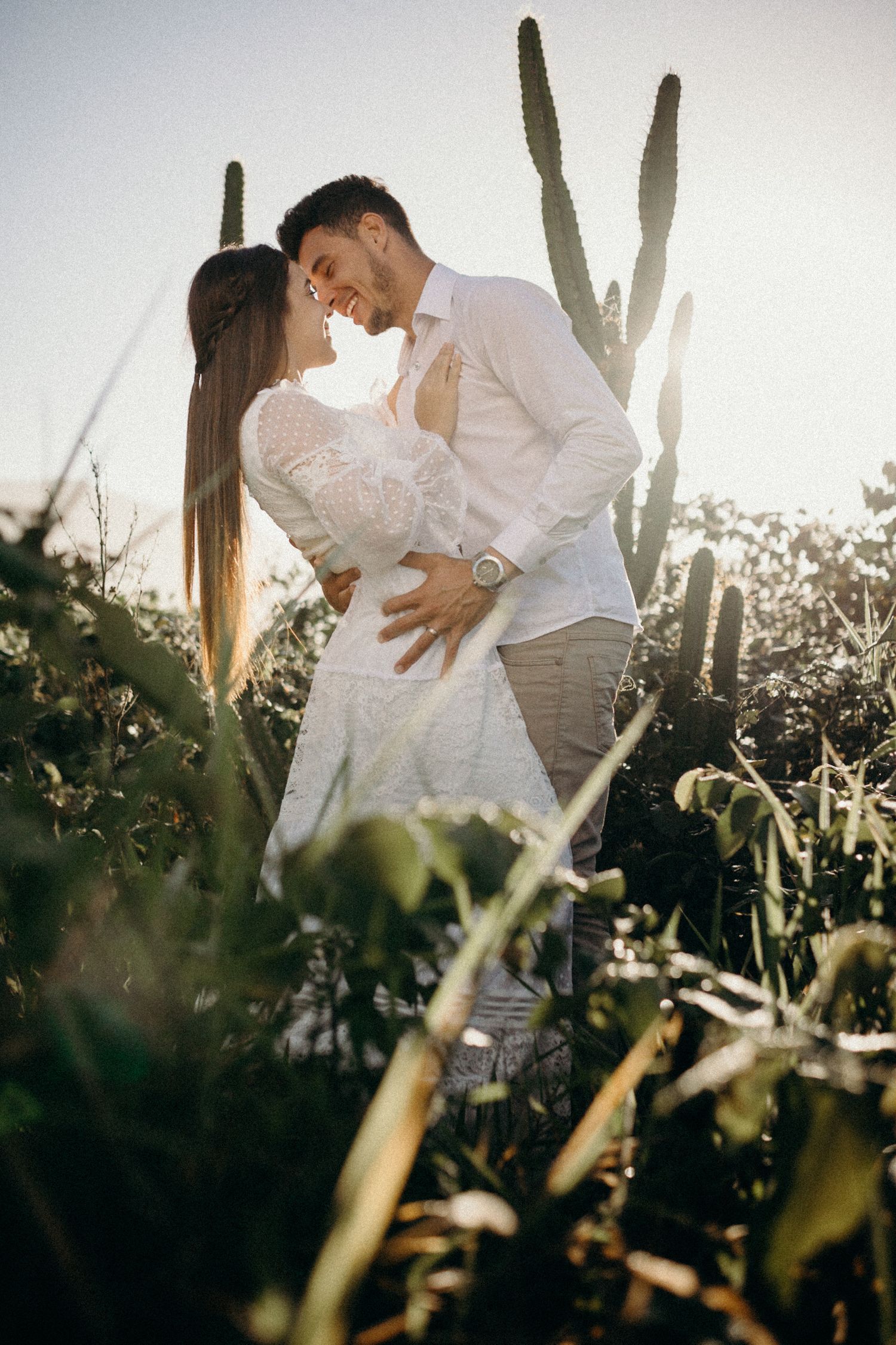 Intimate couple poses against tall desert cactus with sunlight filtering through at sunset.