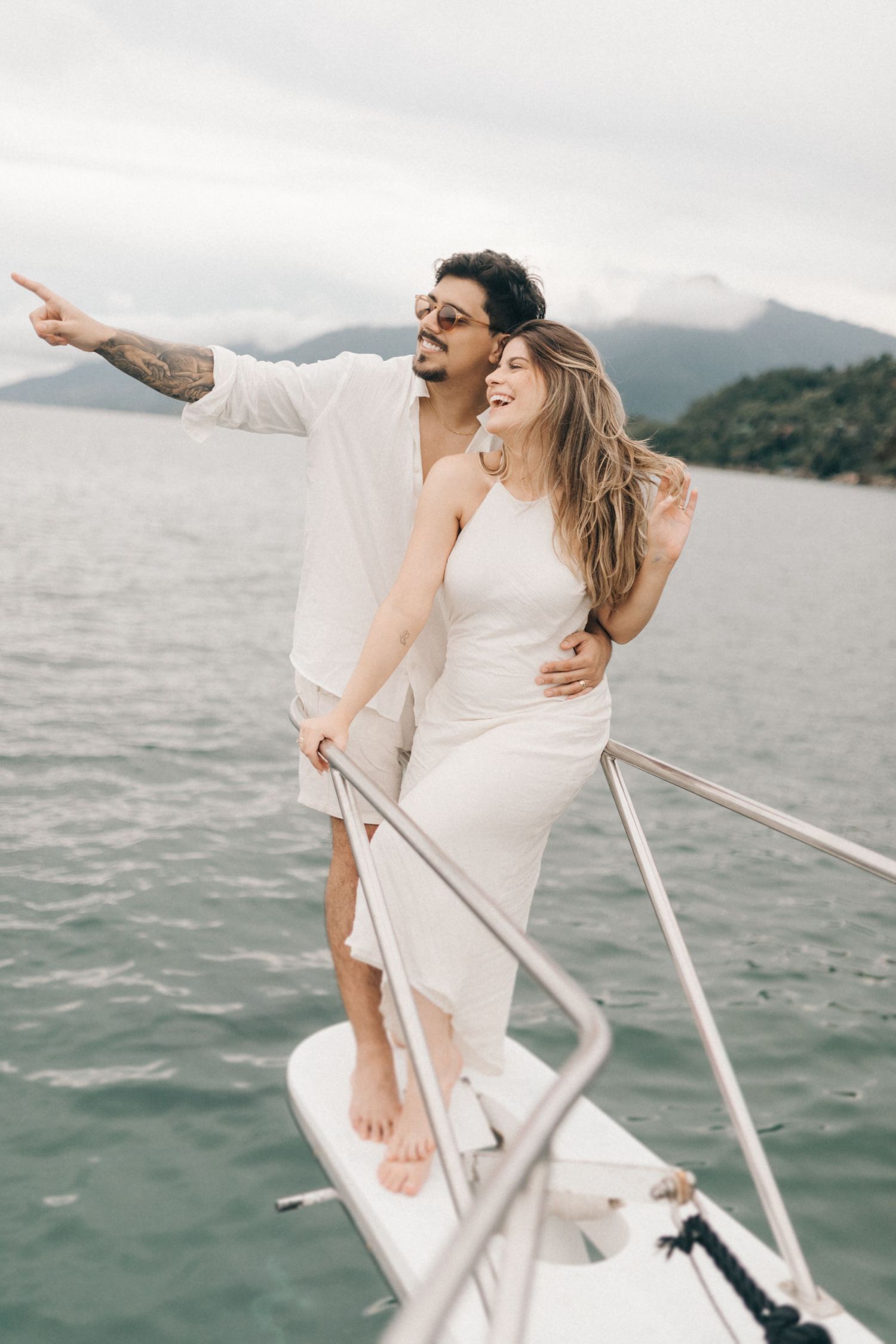 Couple in white clothing standing on a boat bow with mountains and ocean in background.
