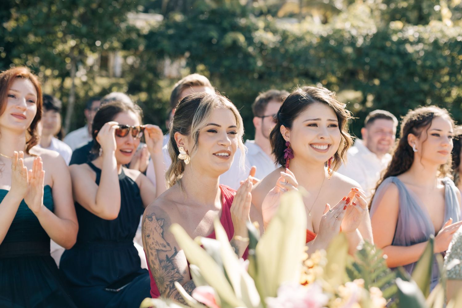 Wedding guests in navy blue dresses clap and smile during an outdoor ceremony.