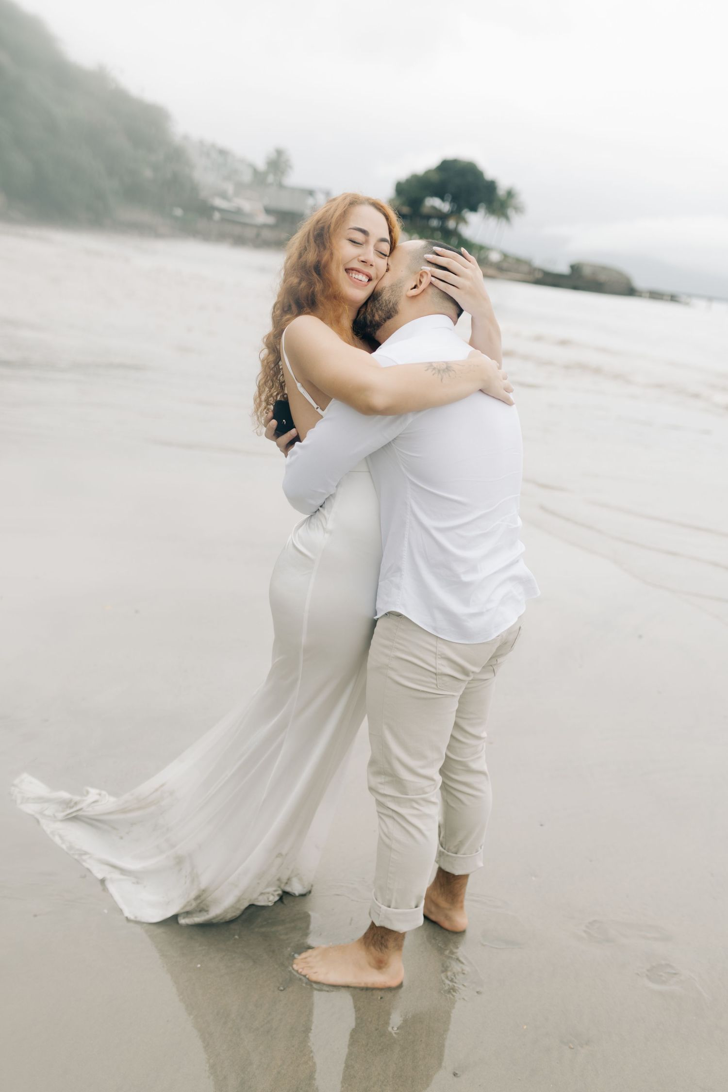 Two people share a joyful embrace on a misty beach, wearing white and creating reflections in the wet sand.