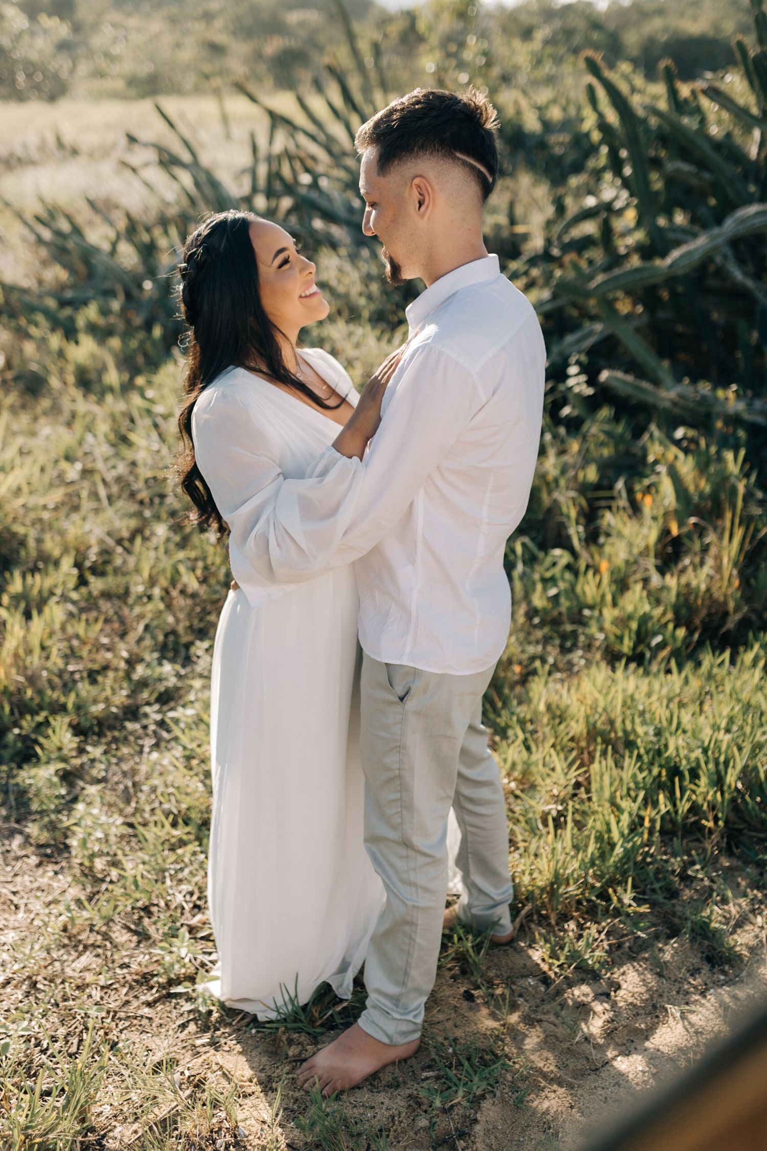 A romantic couple in white clothing embraces while standing on a nature trail at sunset.