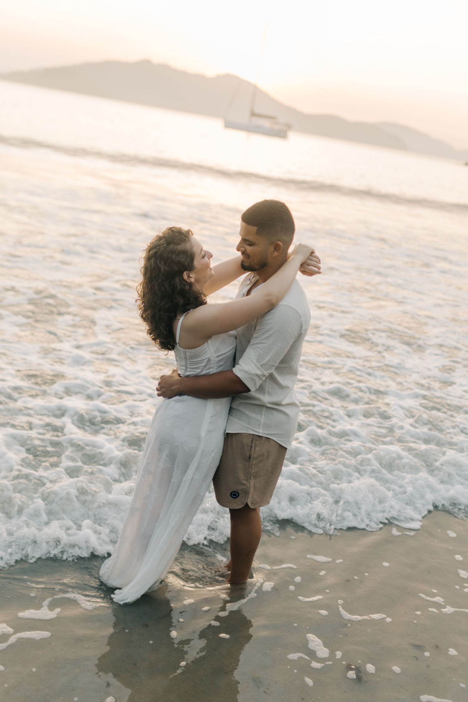 Couple embracing in shallow ocean waves during golden hour with mountains in background.