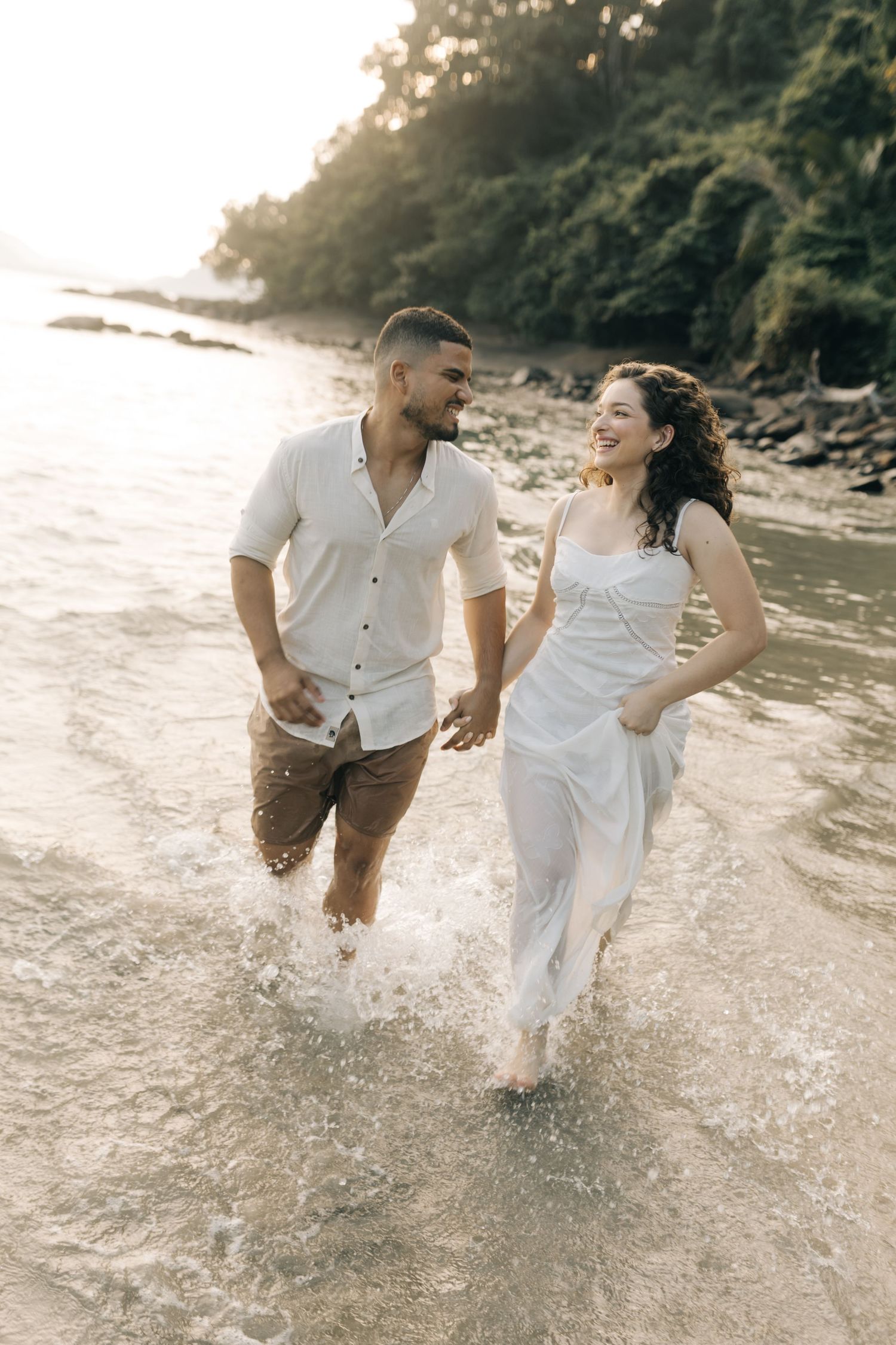 Joyful couple running through shallow ocean water in white outfits.