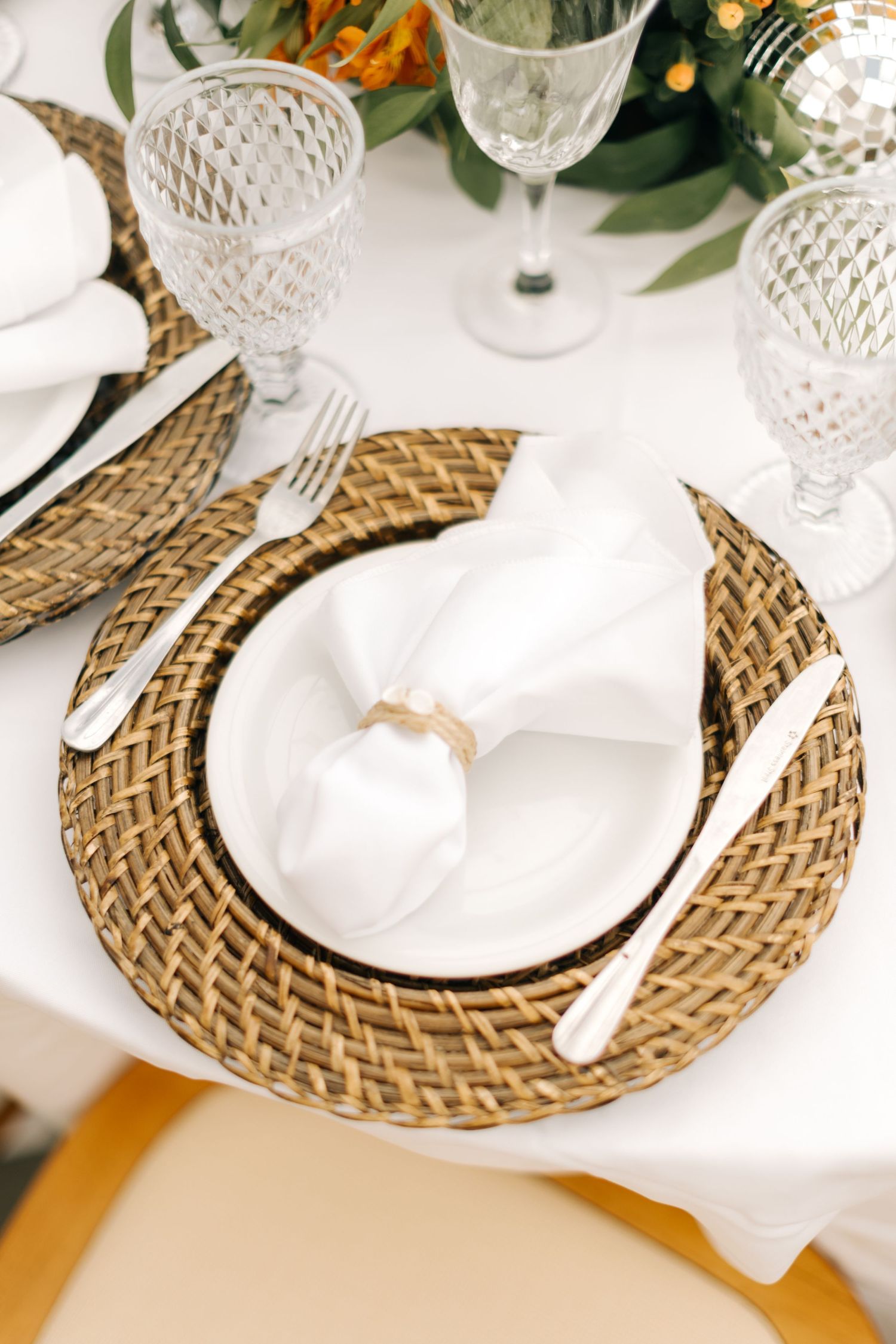 Place settings with woven chargers, white napkins, and crystal glassware on dining table.