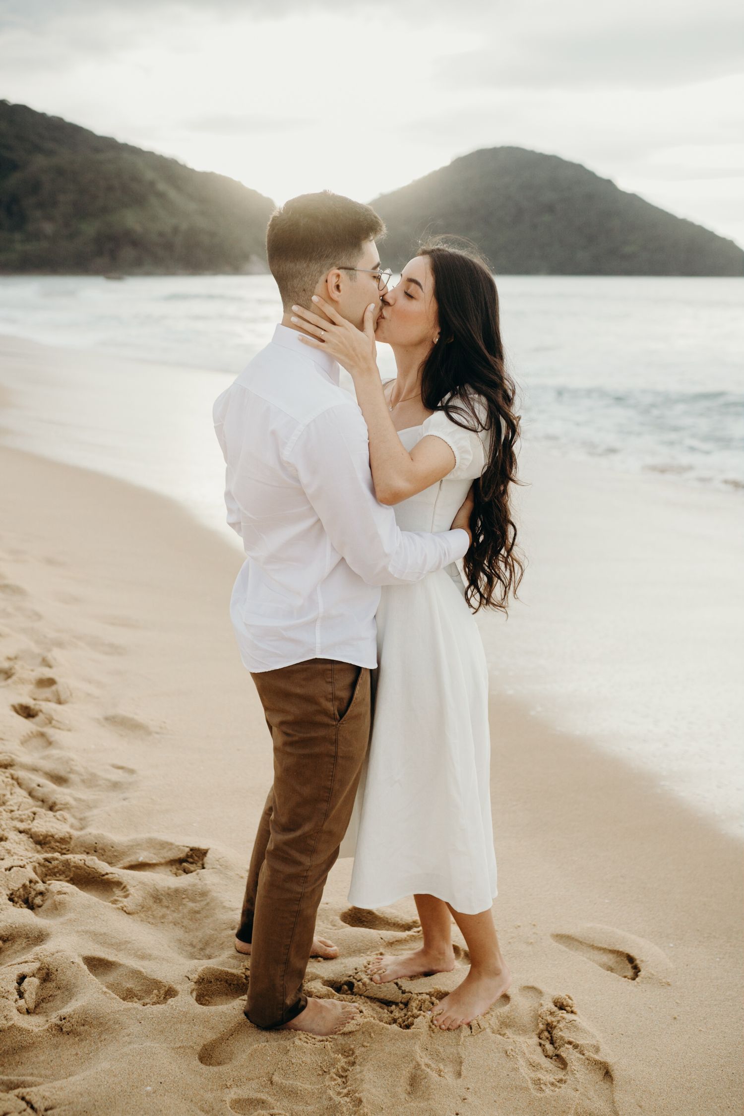 A romantic kiss shared between a couple on a sandy beach with mountains in the background.