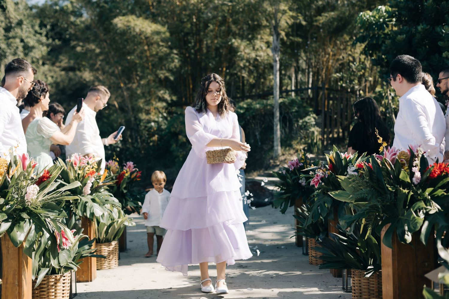 A person in a white dress stands between rows of potted tropical plants during an outdoor ceremony.