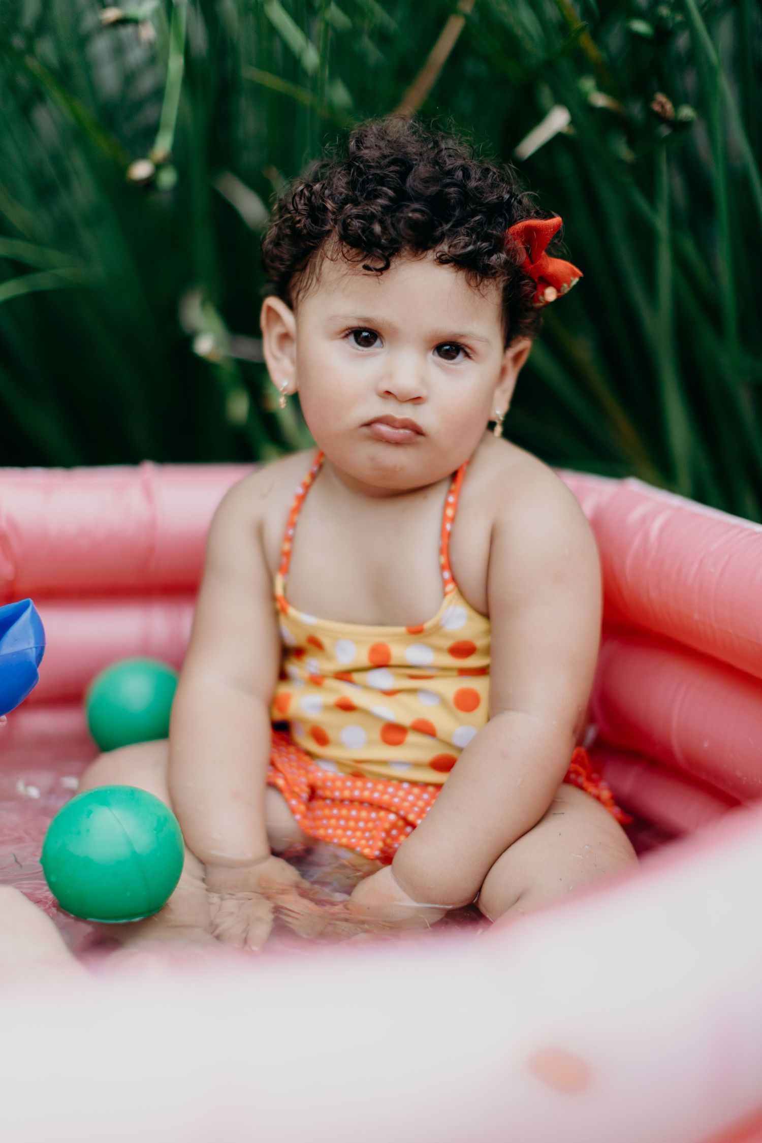 Small child wearing orange polka dot swimwear sits in a pink inflatable pool with green balls.