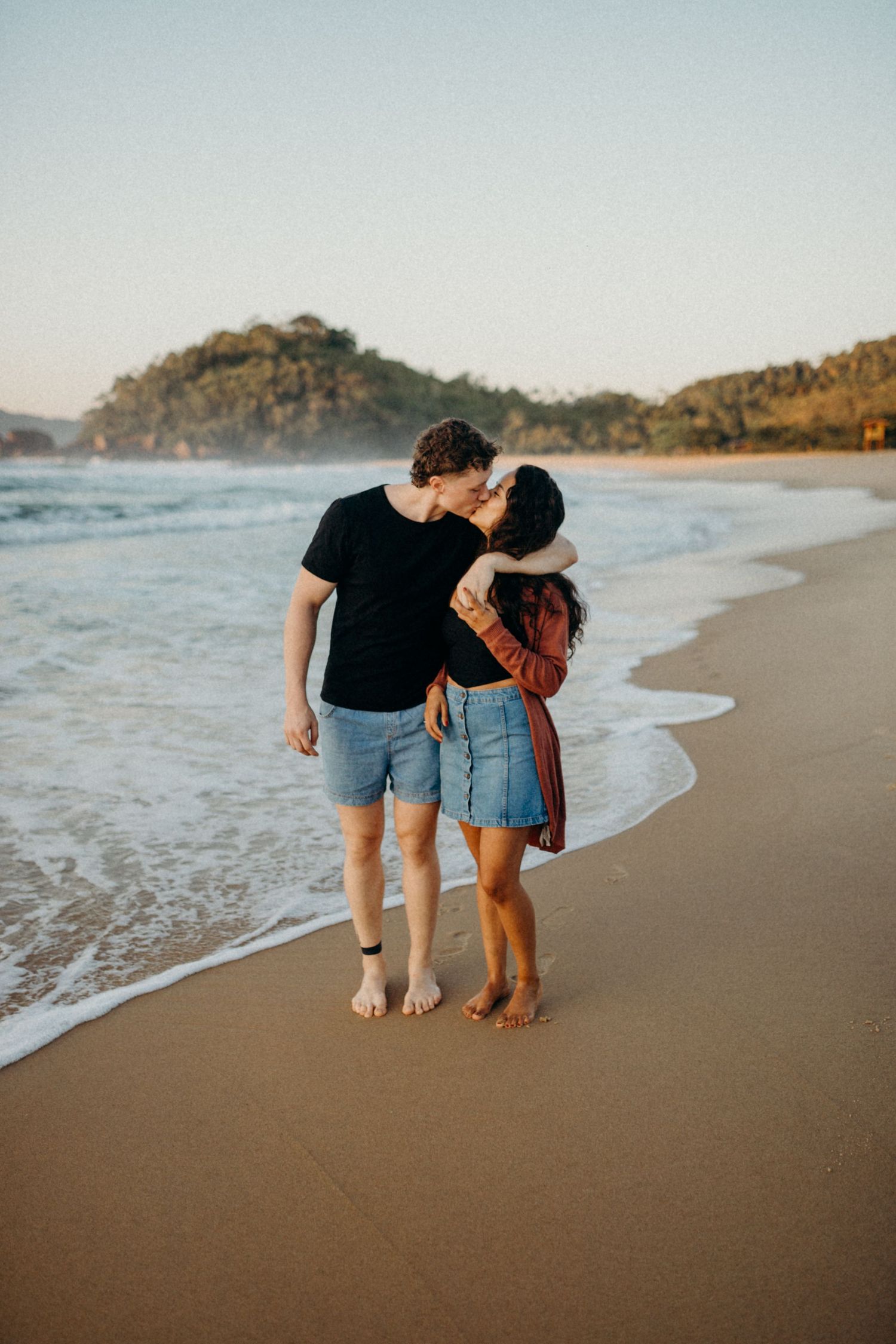 Intimate moment between couple walking on beach with waves and mountains in golden hour light.