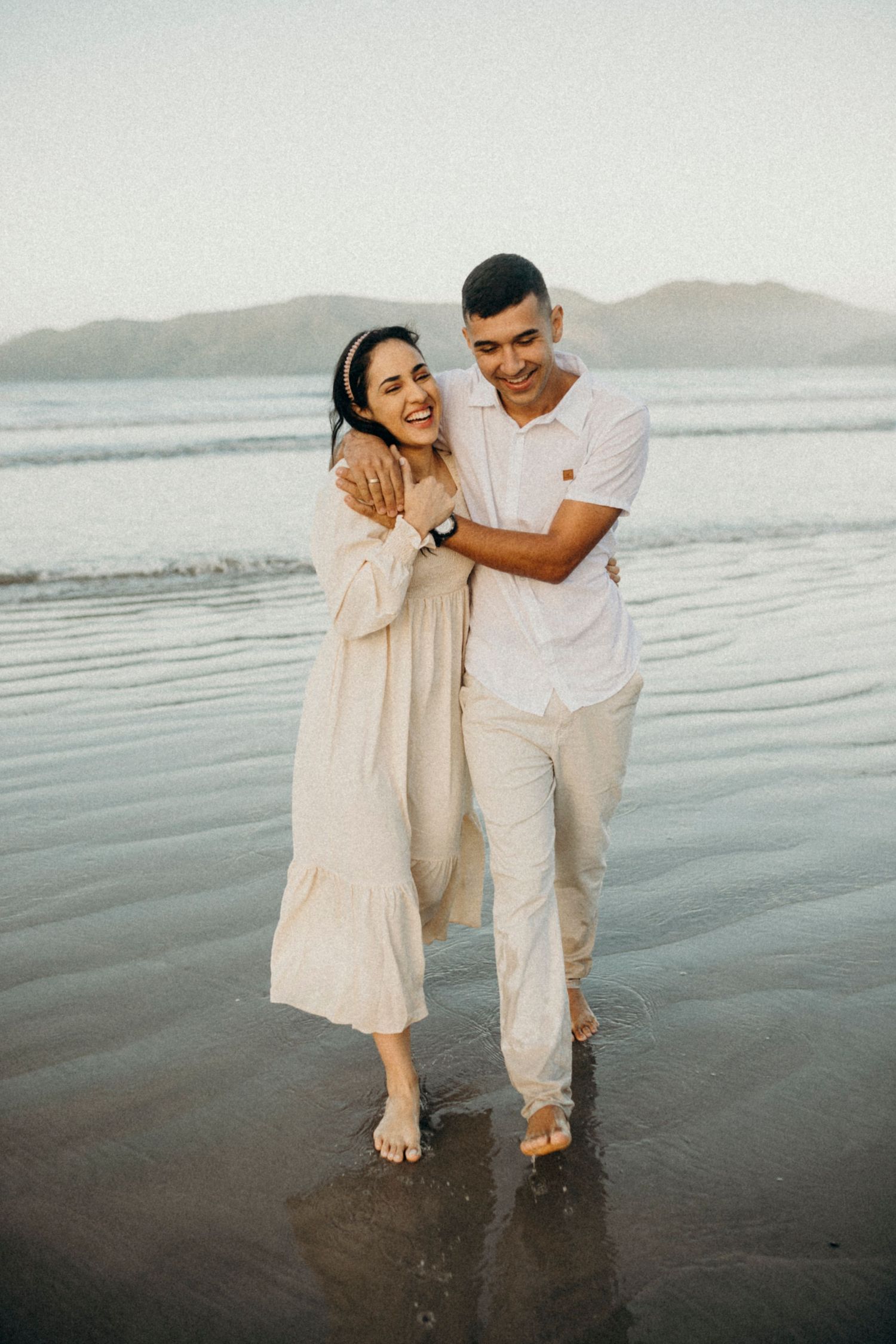 Two people in light colored clothing laugh and play together in the shallow ocean water.