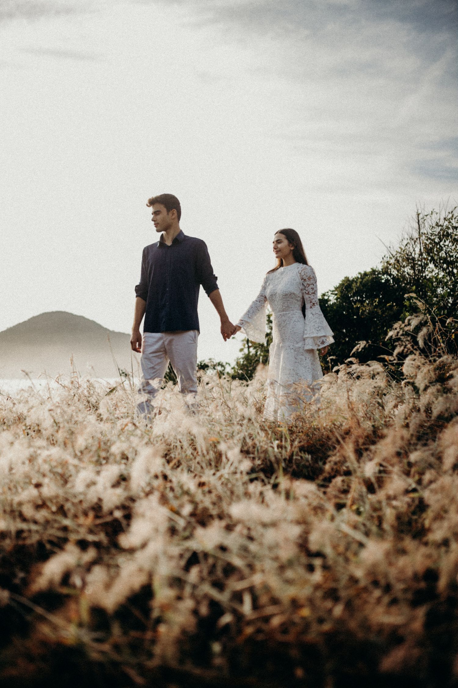 A couple walks through a field of tall grass with mountains in the distance.