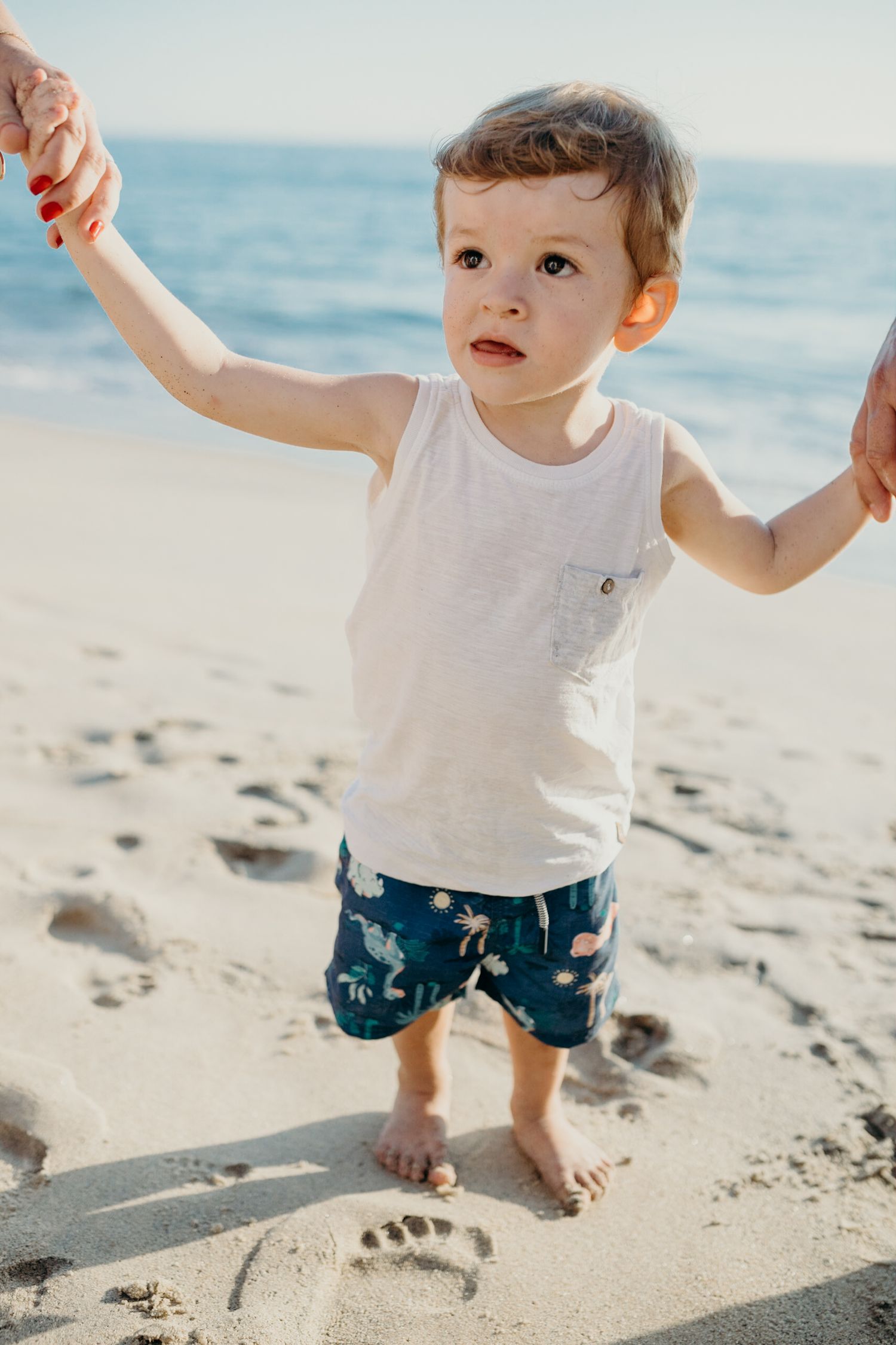 Small barefoot toddler wearing white shirt and navy shorts plays in beach sand during sunny day.