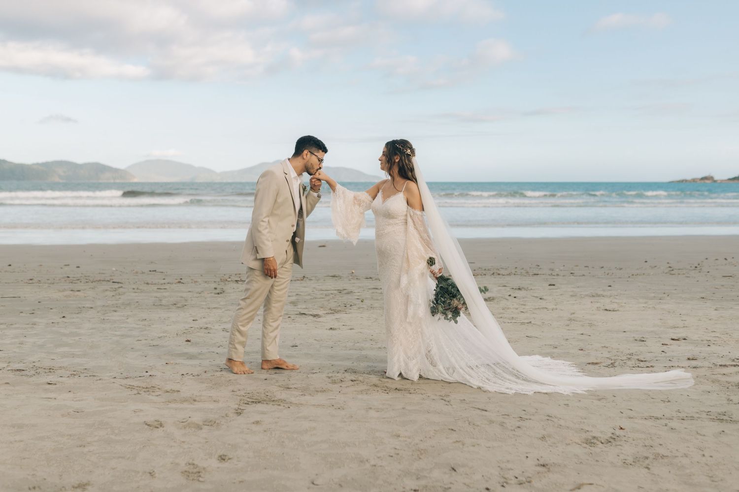 Couple in wedding attire share a playful moment on a scenic beach with mountains in the background.