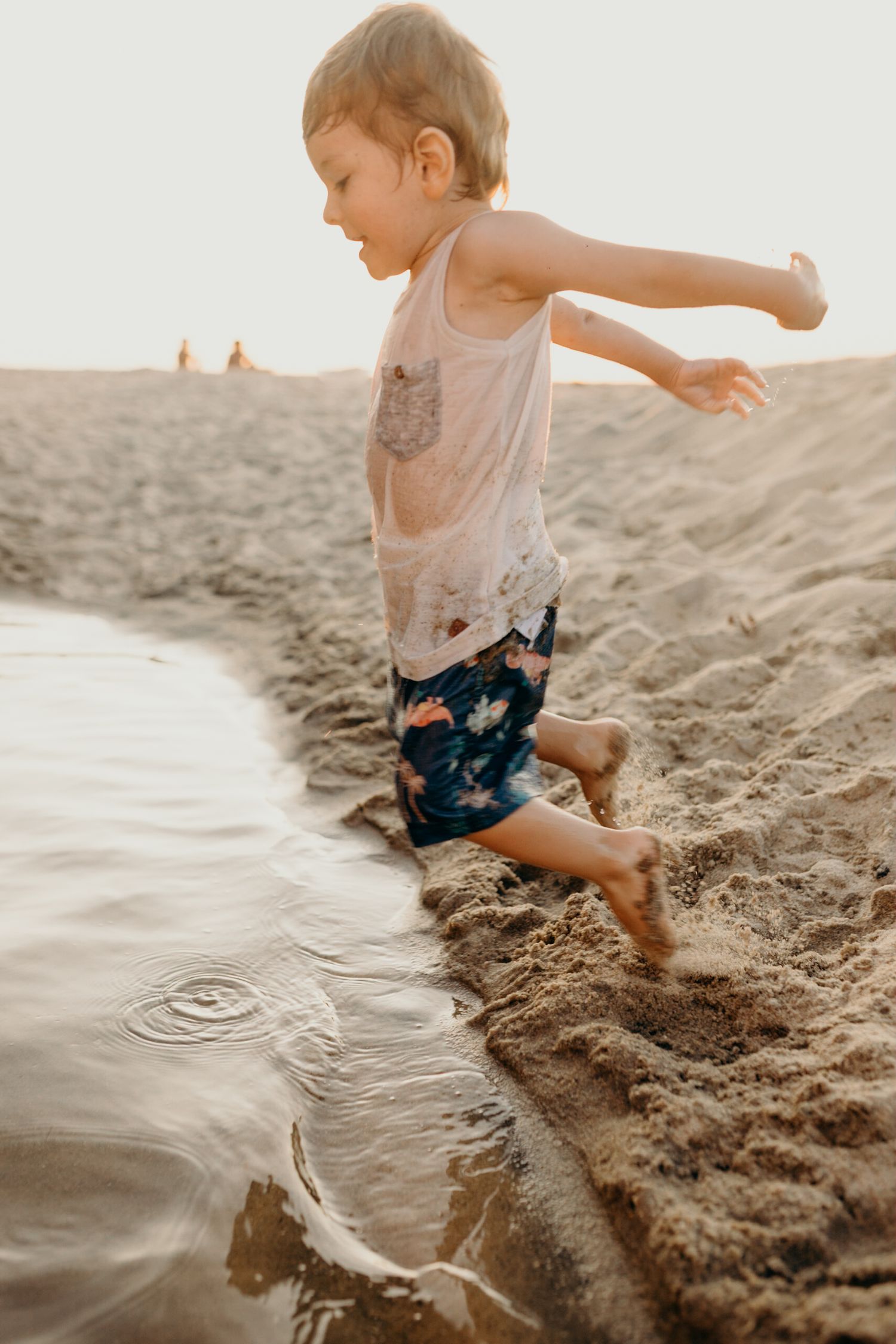 Child jumping into shallow beach water while wearing wet clothes at sunset.