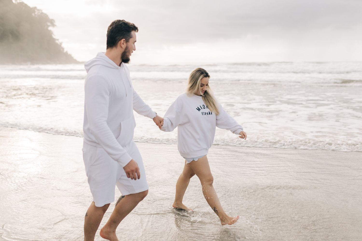 A couple in white outfits runs playfully along a misty beach at sunrise.