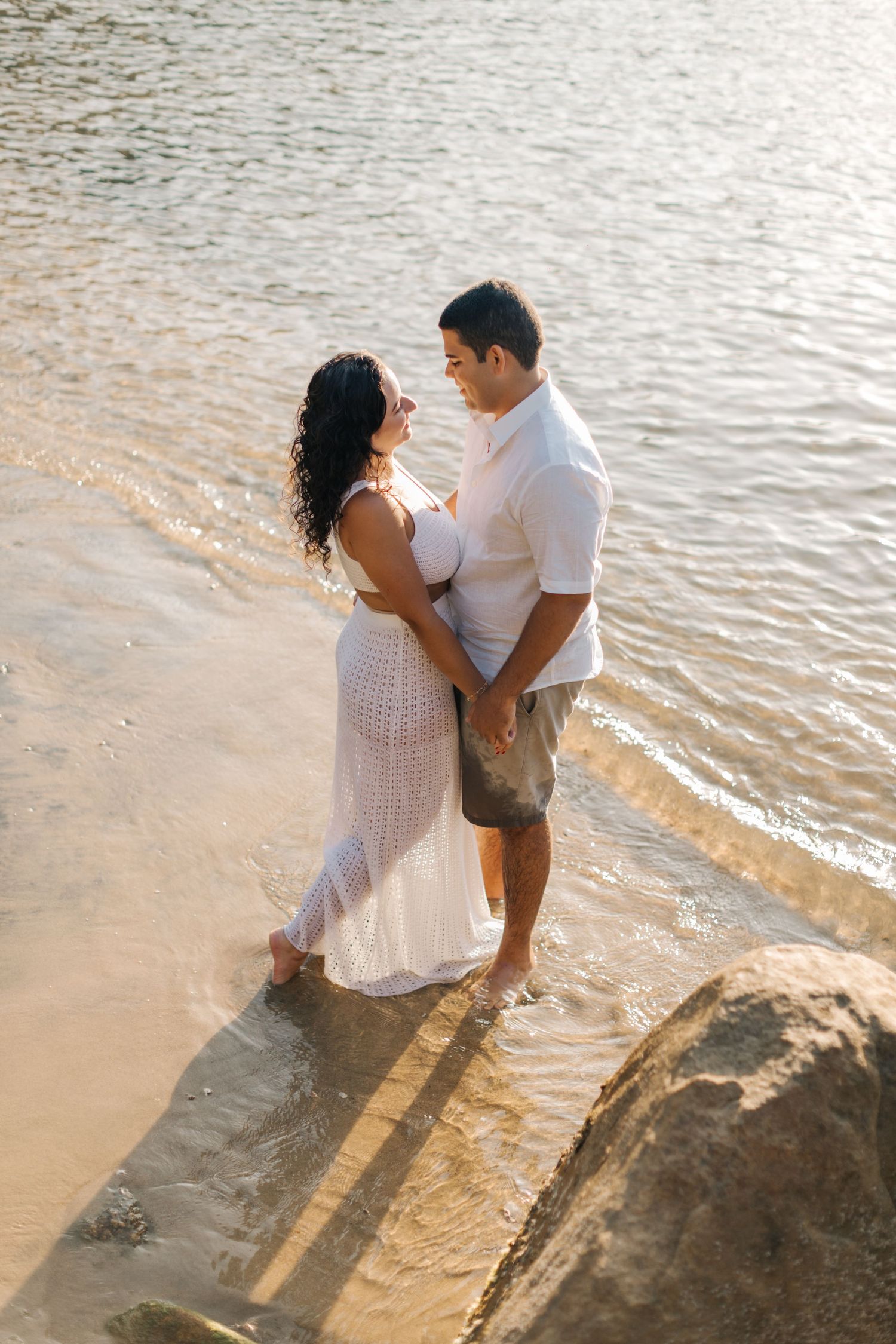 Couple in white clothing embraces at water's edge during golden hour.
