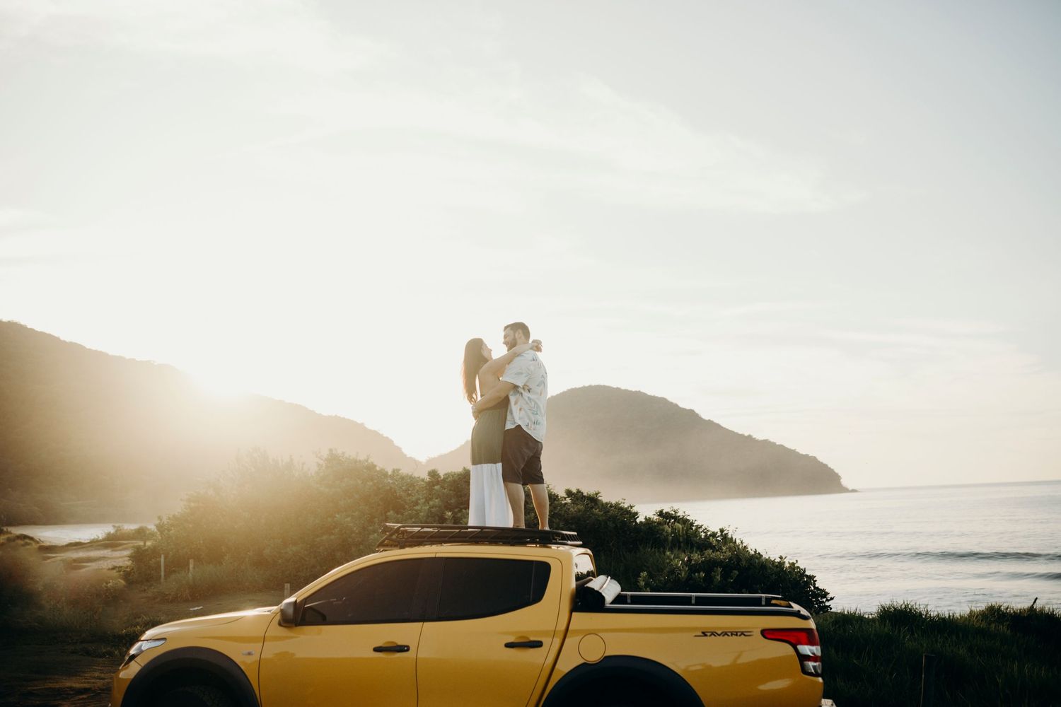 Yellow pickup truck parked at sunset with silhouettes against a coastal mountain backdrop.
