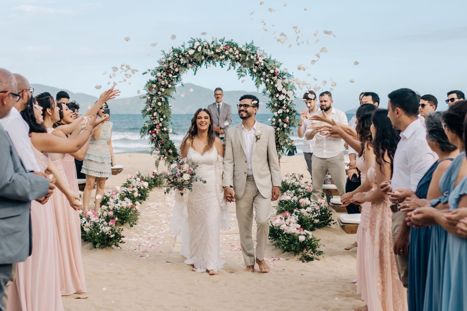 Newlyweds walk down the beach aisle under a floral arch as guests throw petals.
