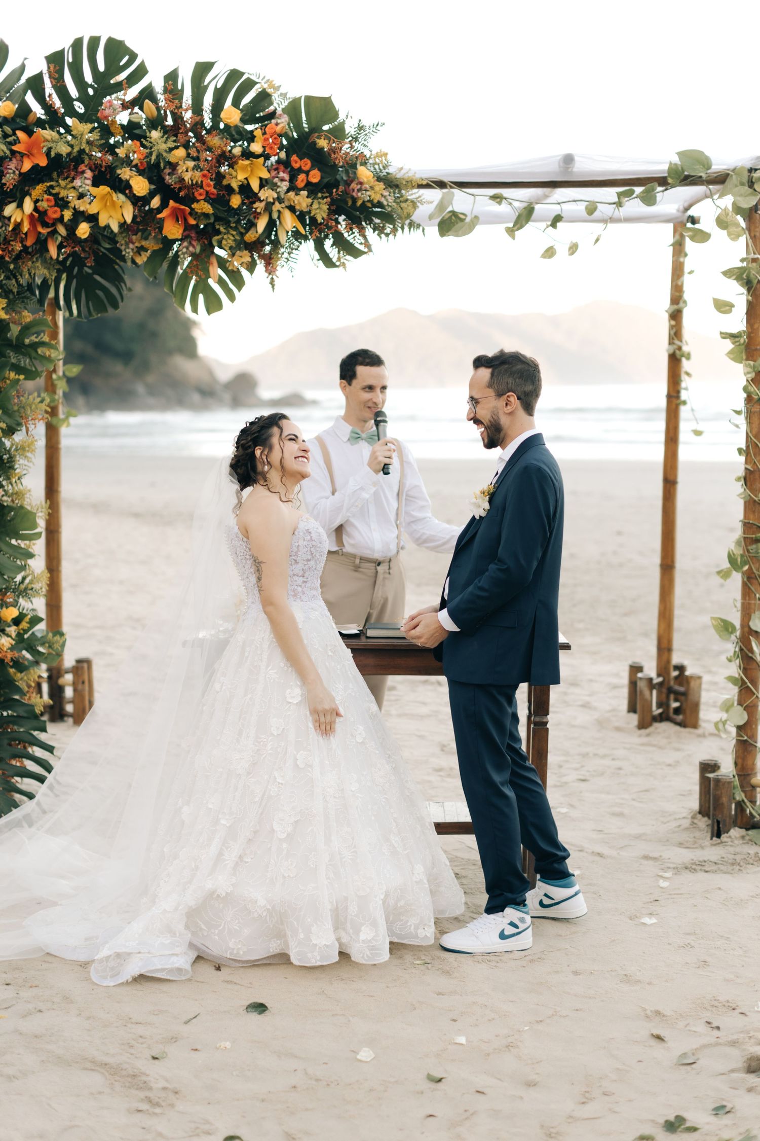 Joyful wedding moment under a tropical floral arch on a sandy beach with mountains in the background.