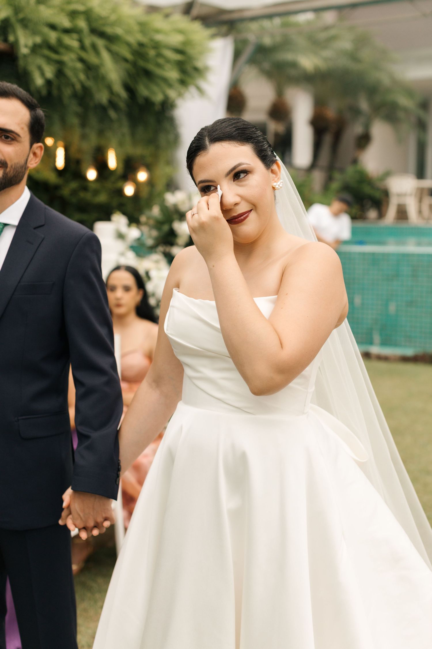 A joyful wedding couple outdoors with the bride in a white strapless gown wiping away a happy tear.