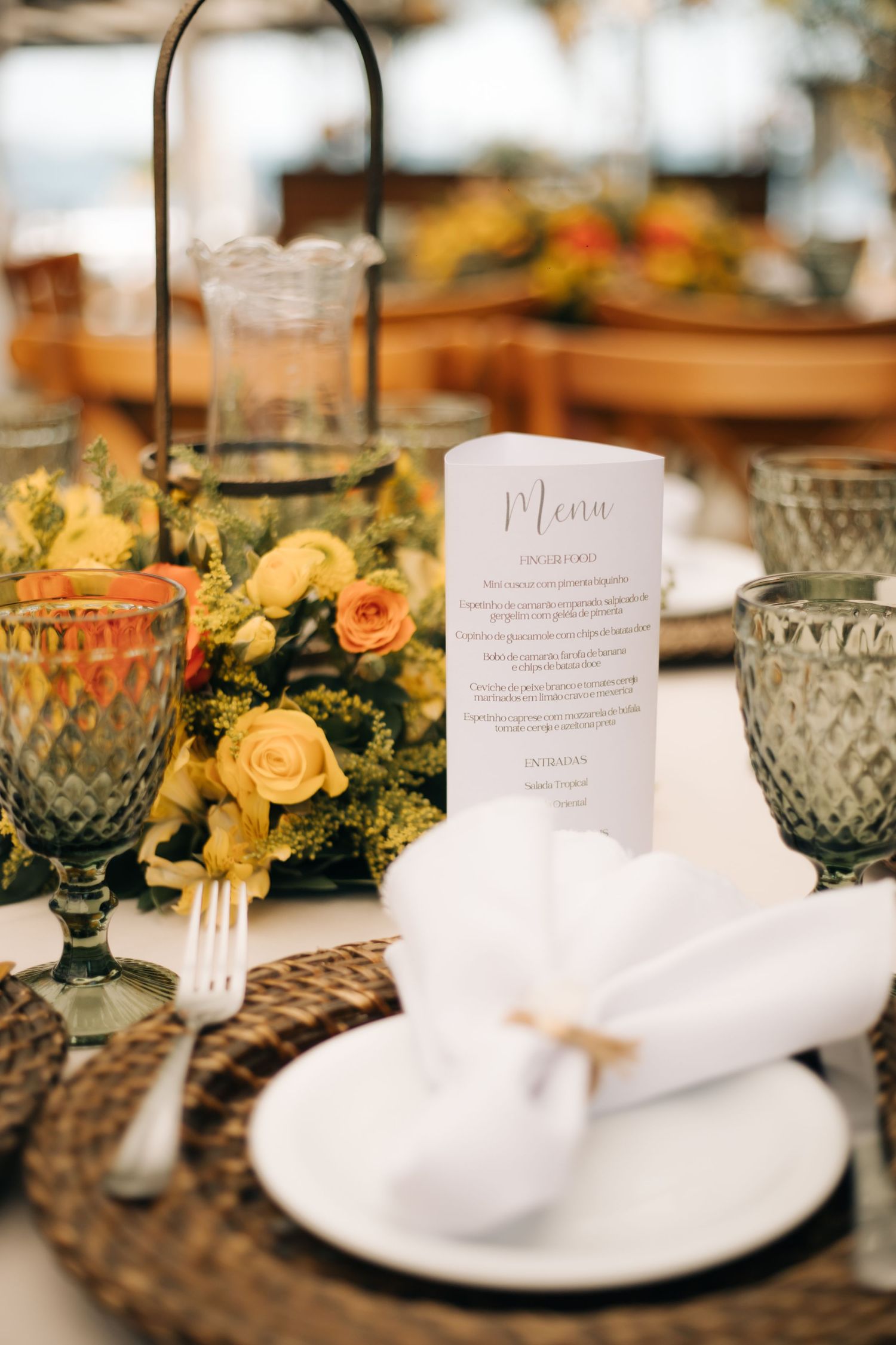 Wedding table setting with yellow roses, crystal glasses, and white menu cards on woven placemats.