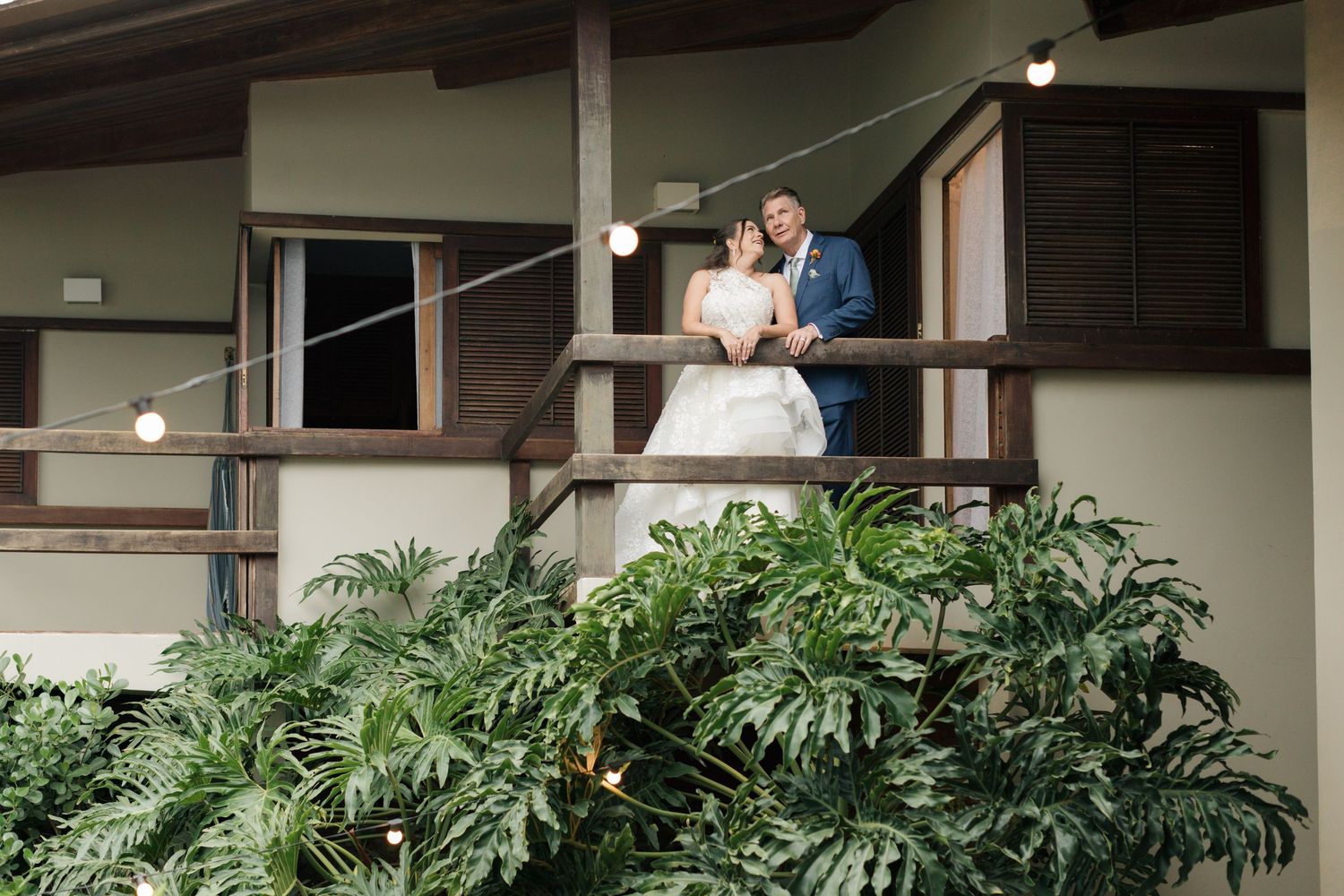 A couple embraces on a covered patio with lush monstera plants creating a tropical atmosphere.