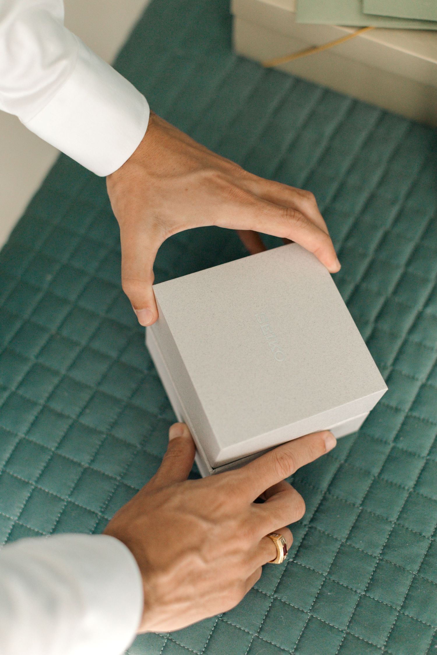 Hands holding a light gray luxury gift box on a quilted emerald green surface.