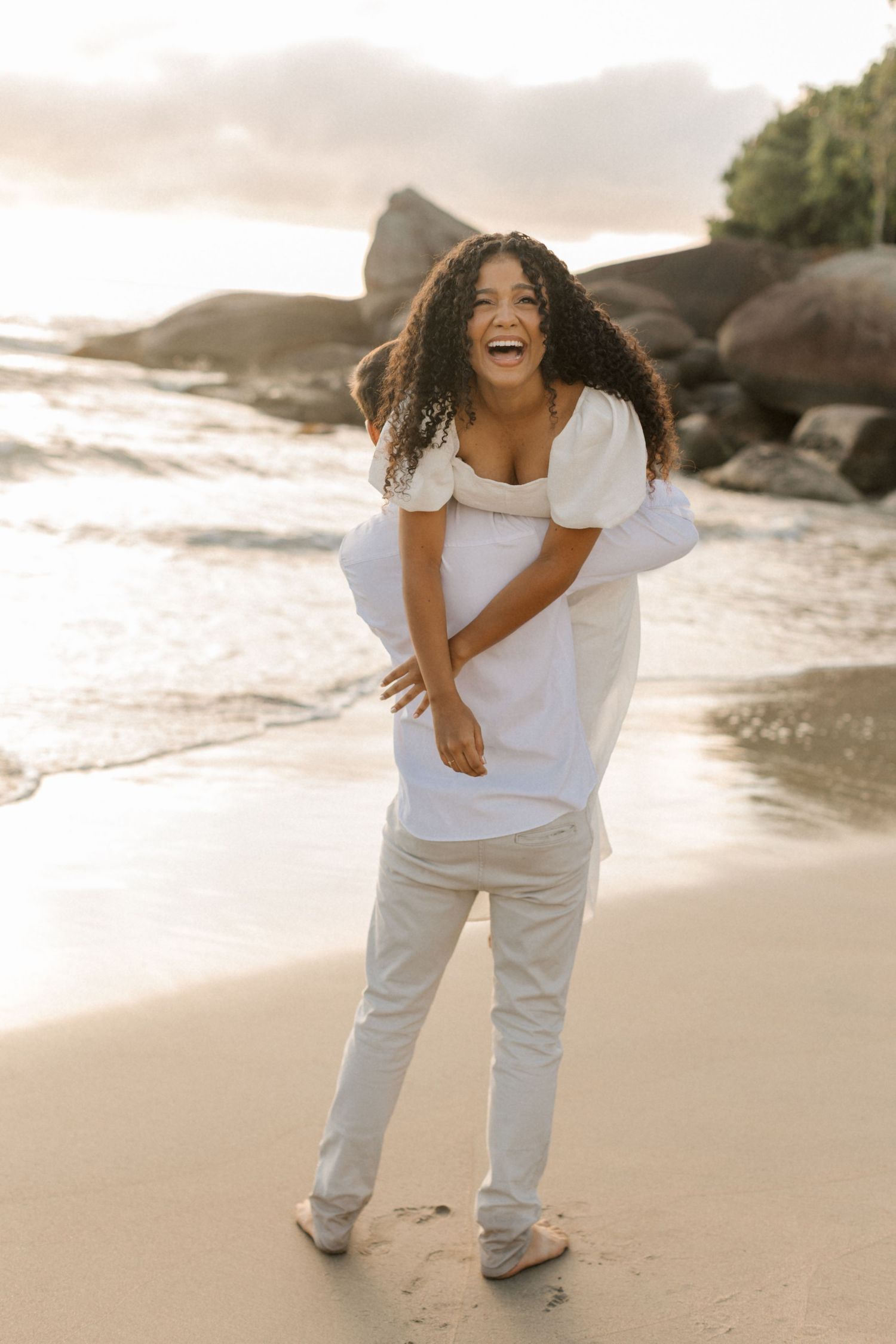 A laughing person wearing a white outfit enjoys a warm sunset moment on a rocky beach with waves crashing behind them.