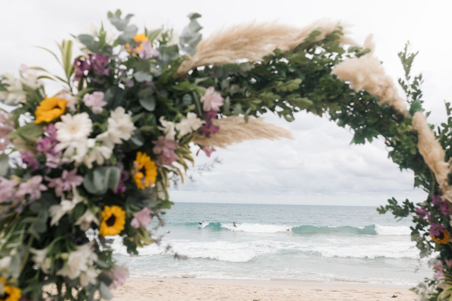 Rustic beach wedding arch adorned with wildflowers and pampas grass against ocean backdrop.