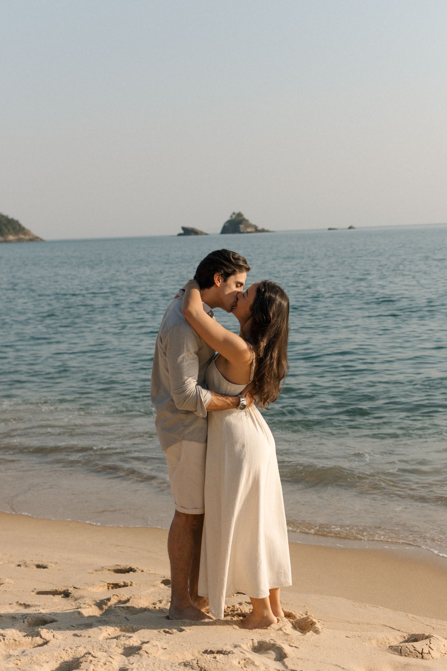 A romantic moment unfolds as a couple embraces on a tranquil beach with ocean waves and rocky outcrops in the distance.