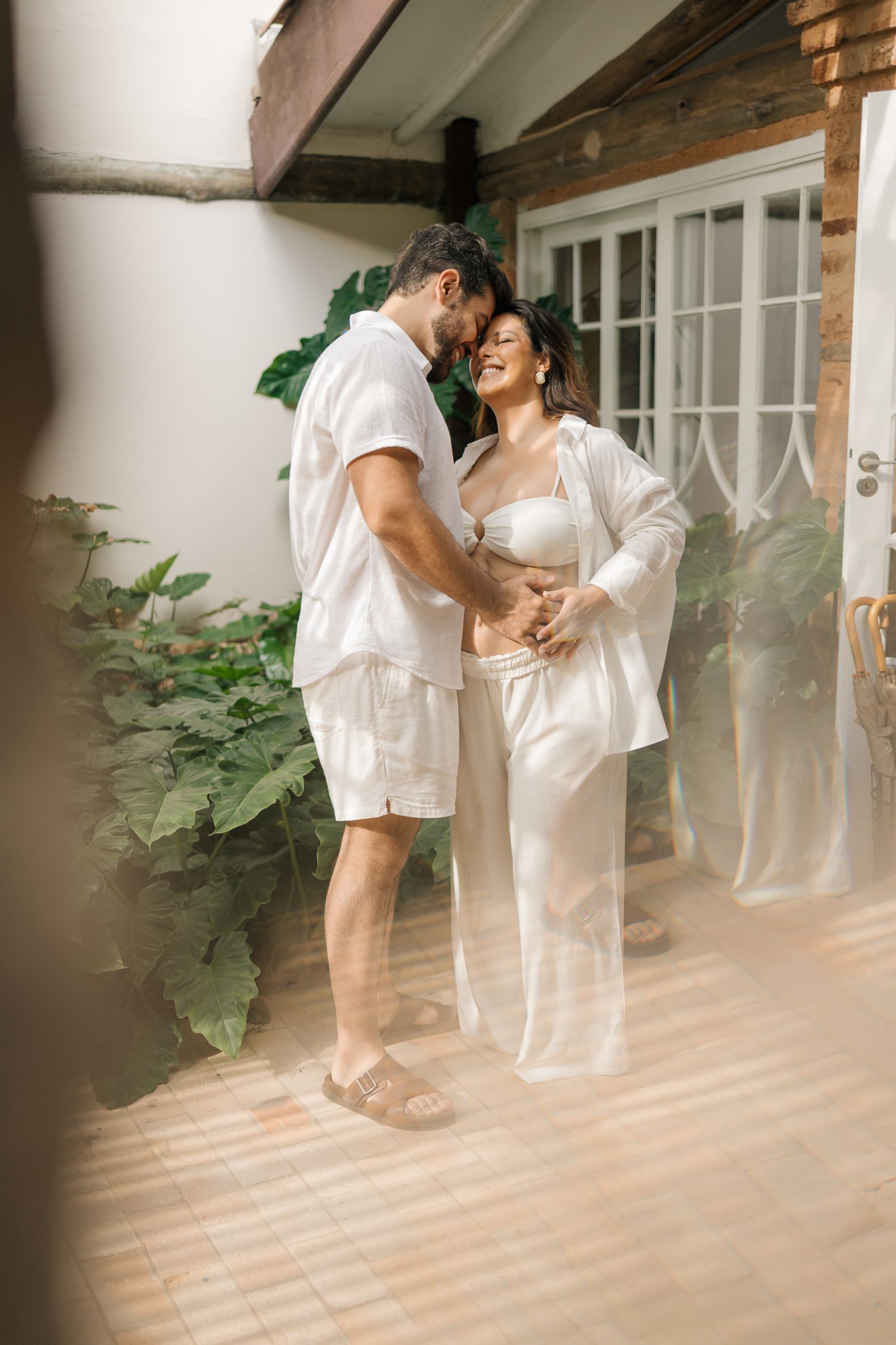 A couple in white outfits embraces tenderly in a sunlit room with plants and rustic wooden beams.