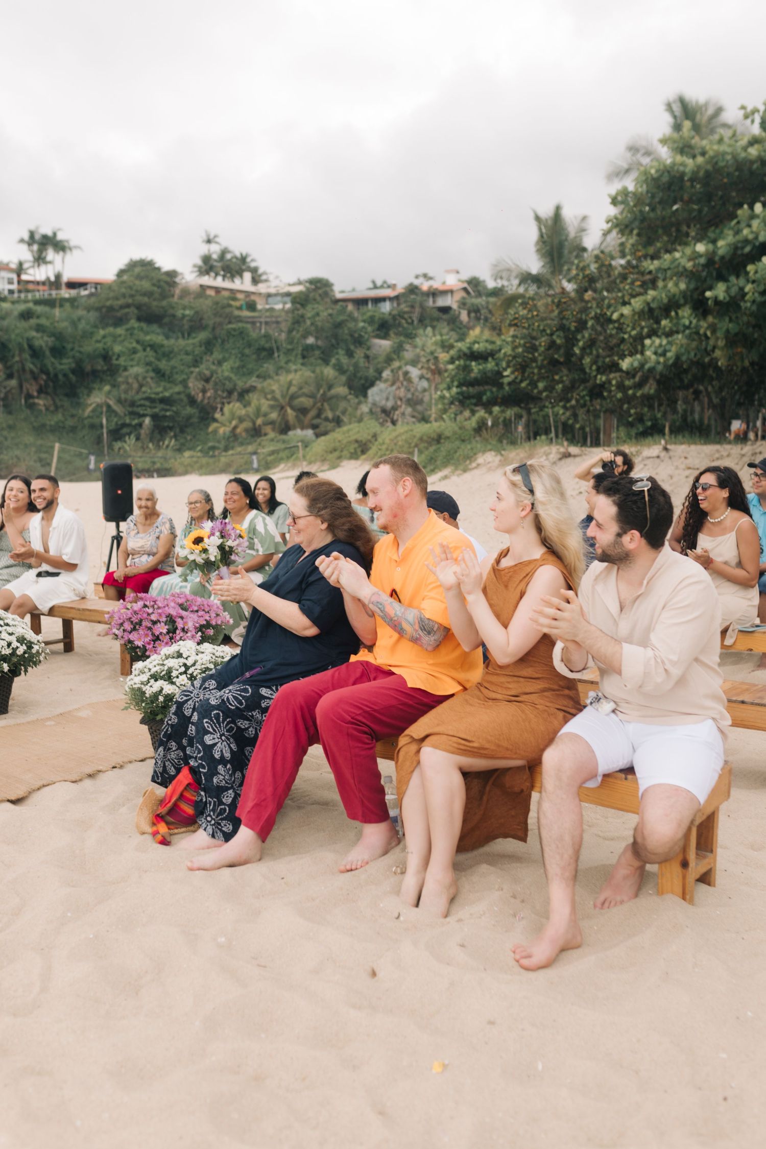Wedding guests seated on wooden chairs at a beachside ceremony with pink floral decorations.